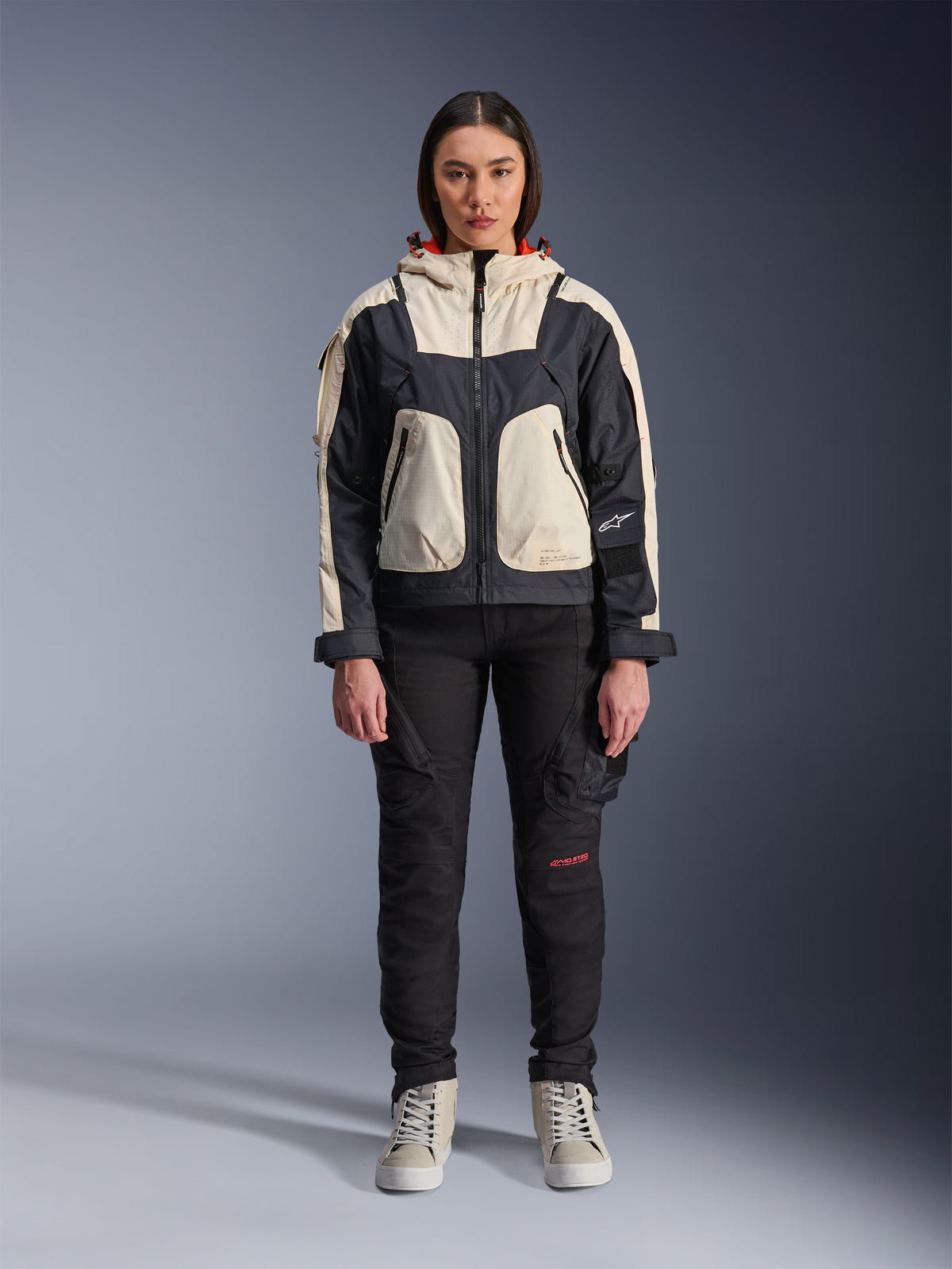 Kvinnlig modell som bär Alpinestars Molly Air-jacka i Black Almond, med en färgblockerad design med krämfärgade och svarta paneler och en huva med rött foder, matchad med svarta motorcykelbyxor och krämfärgade sneakers, studiobild av urban samtida körutrustning.