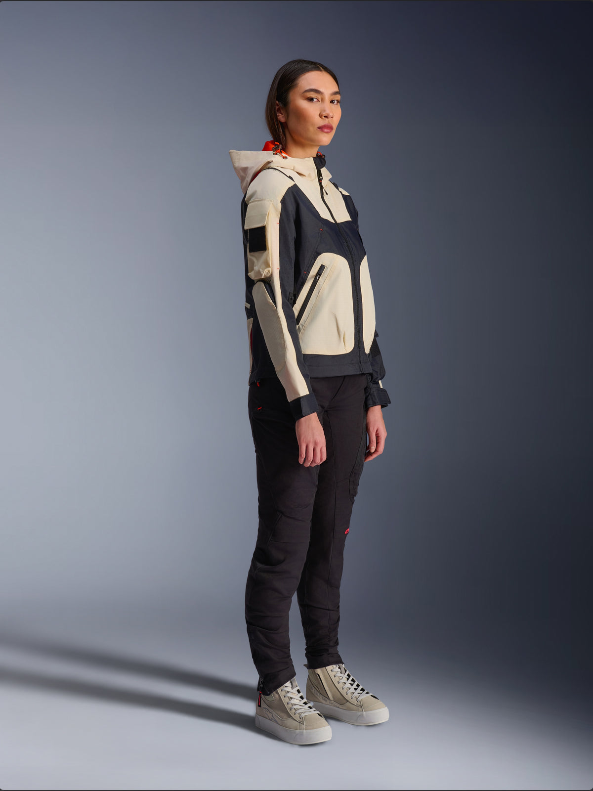 Alpinestars Molly Air-jacka i Black Almond, bärs av en kvinnlig förare med svarta byxor och krämvita high-top sneakers, med en tvåfärgad design med svarta och beigea paneler, integrerad huva och modern urban stil.