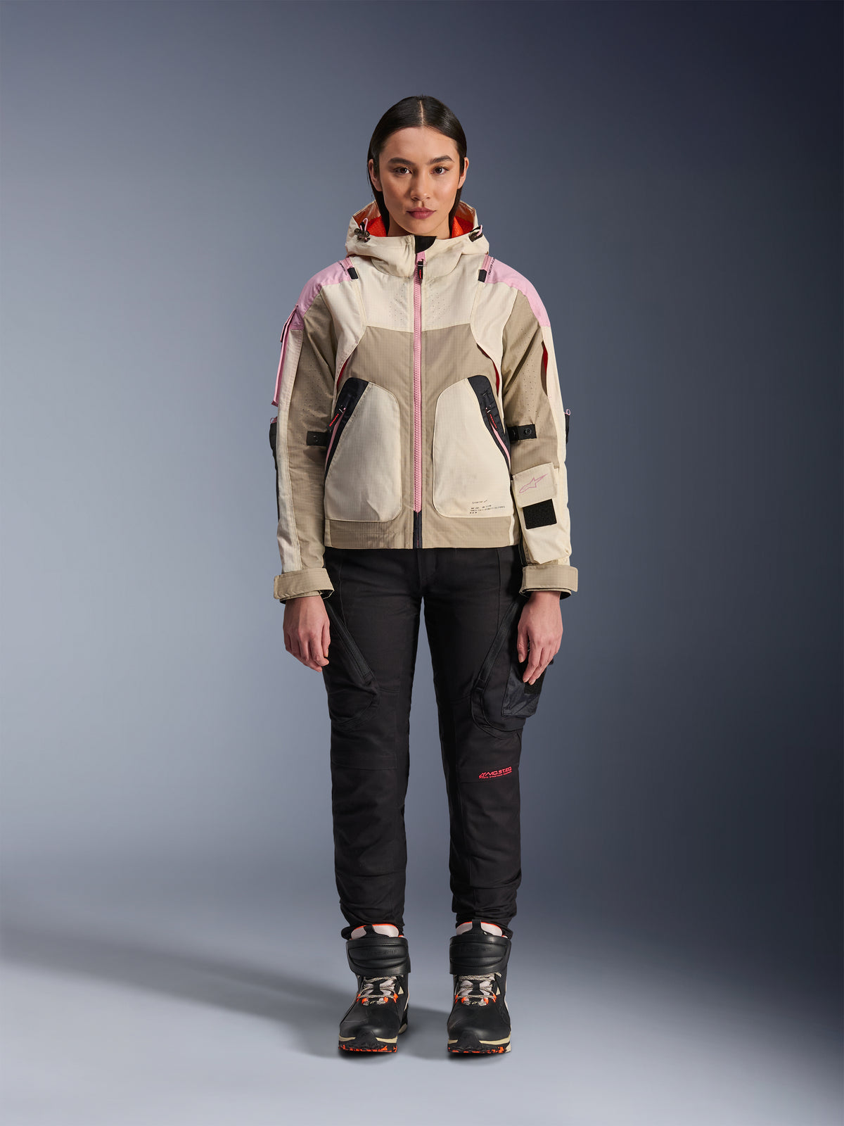 Kvinnlig modell bär Alpinestars Molly Air Jacka i Sand Pink Almond, kombinerad med svarta tekniska mc-byxor och stövlar, helfigursbild framifrån, urban motorcykeljacka med utility-inspirerad design och rosa detaljer