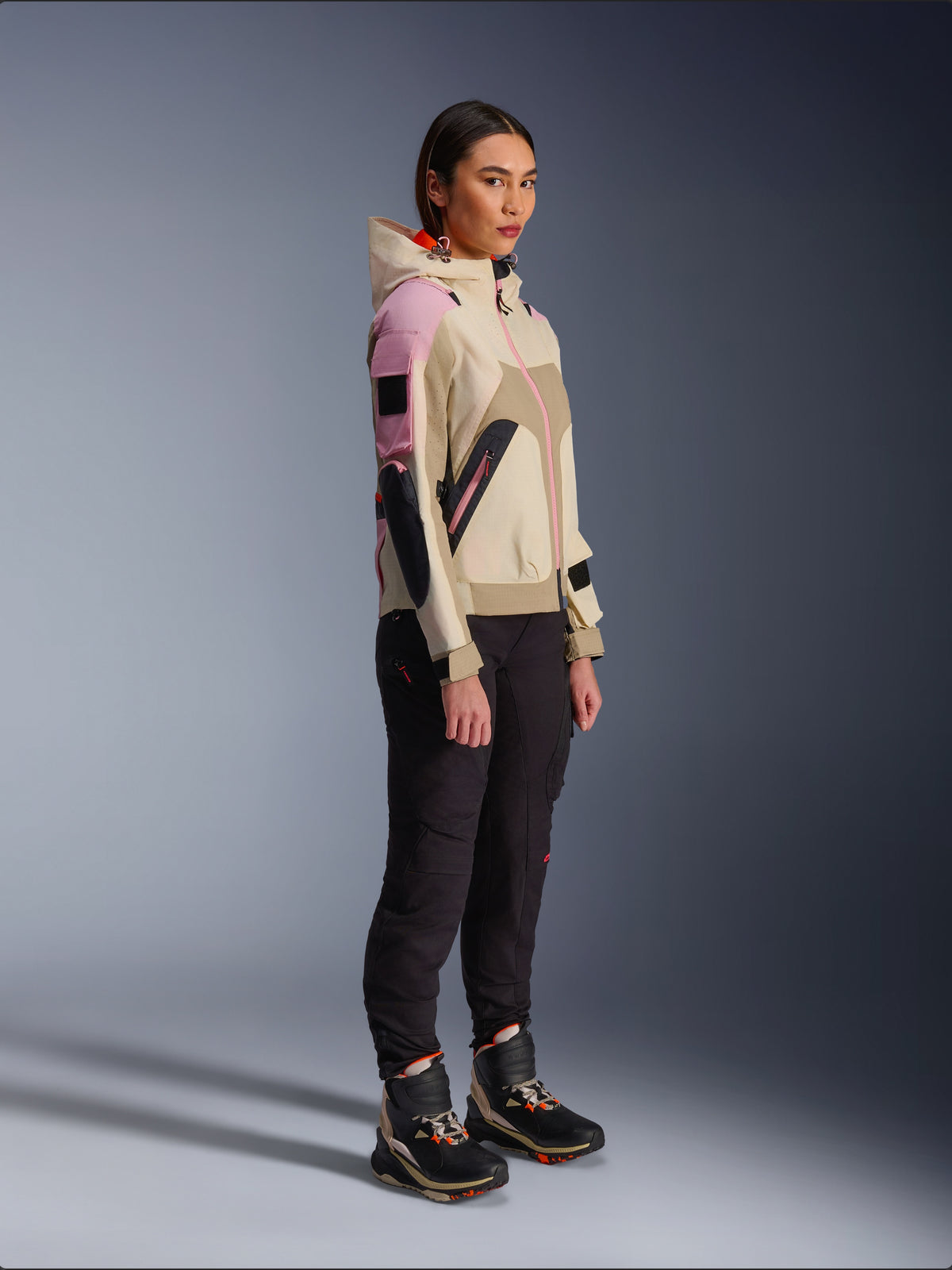 Kvinnlig modell i Alpinestars Molly Air-jacka i Sand Pink Almond, matchad med mörka tekniska byxor och höga körstövlar, urban motorcykeljacka med huva och flerfärgade paneler, samtida streetwear-inspirerad design