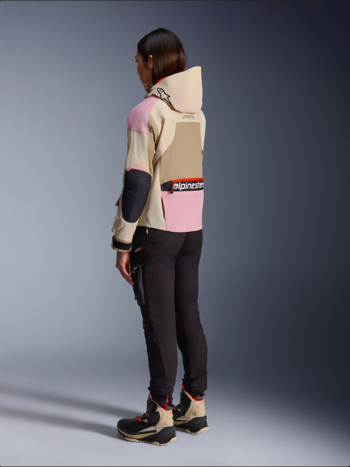 Kvinnlig förare iklädd Alpinestars Molly Air-jacka i Sand Pink Almond, beige och rosa design i flera nyanser, sedd snett bakifrån med svarta mc-byxor och matchande tekniska stövlar, urban motorcykelstil med huva.