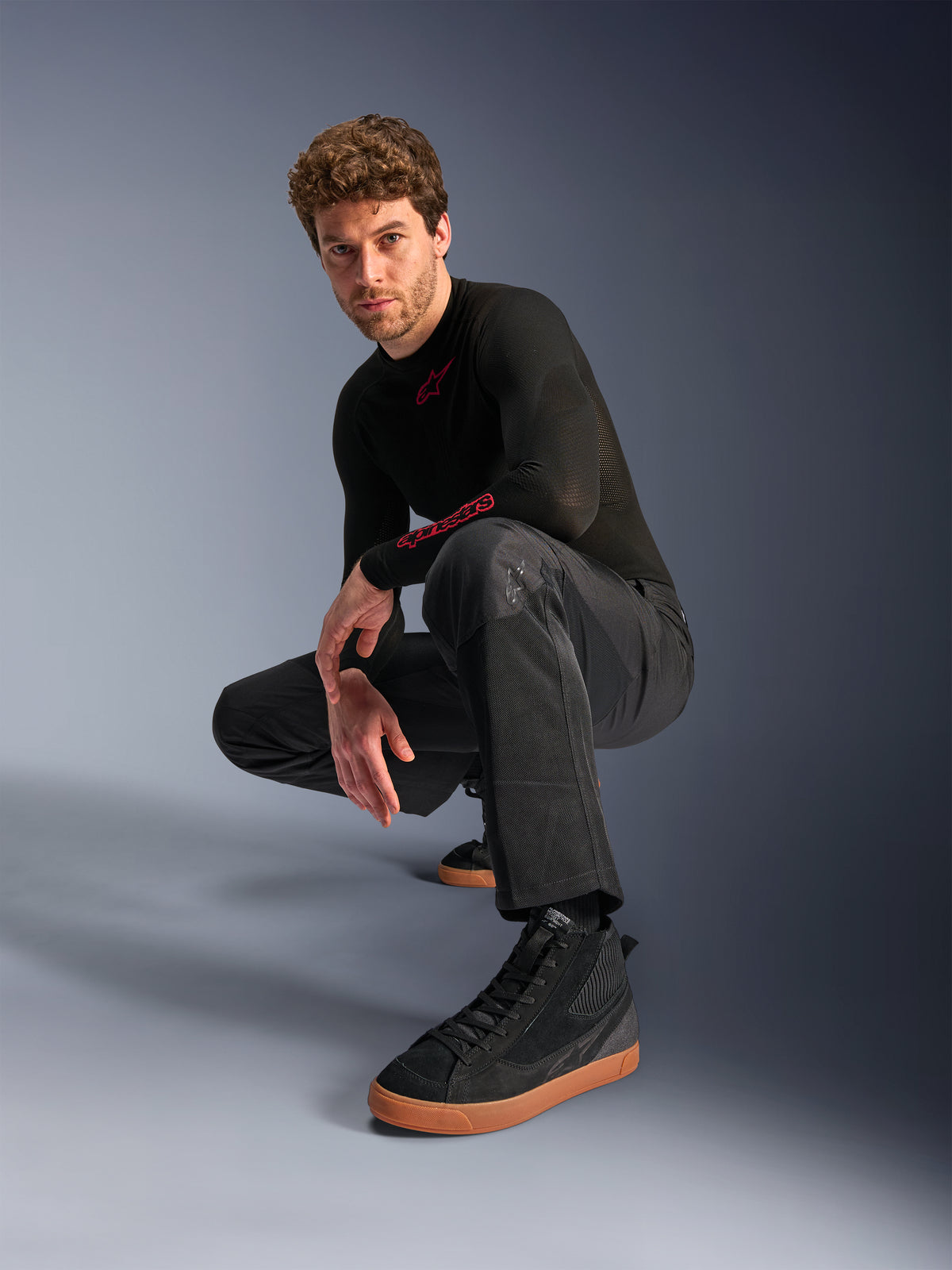 Alpinestars C-1 Air byxor, urbana motorcykelbyxor, svarta, modell som hukar i en studiomiljö, bärs med en svart långärmad tröja och svarta sneakers med bruna sulor, med ventilerande mesh-paneler och en modern urban stil