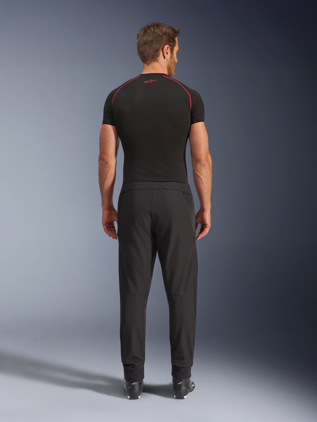 Alpinestars Aeroshell Airflow byxor, svarta motorcykelbyxor, sedda bakifrån på en modell med svart underställ och stövlar, med ett lätt softshell-chassi och integrerade meshpaneler för ventilation.