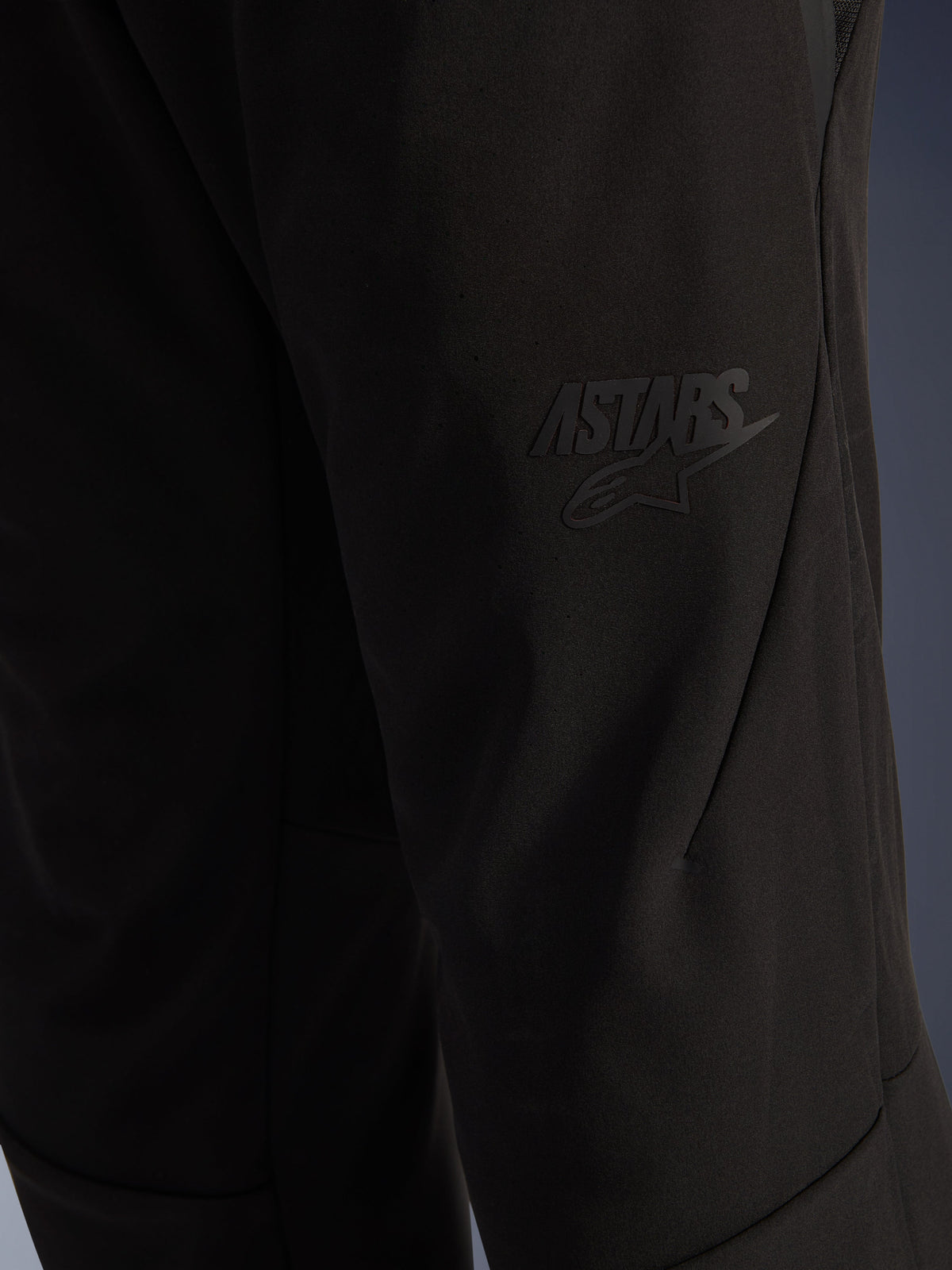 Alpinestars Aeroshell Airflow Pants, svarta motorcykelbyxor, närbild på lårområdet med en upphöjd svart ASTARS-logotyp, lätt stretch-softshell-material med ergonomisk sömdesign