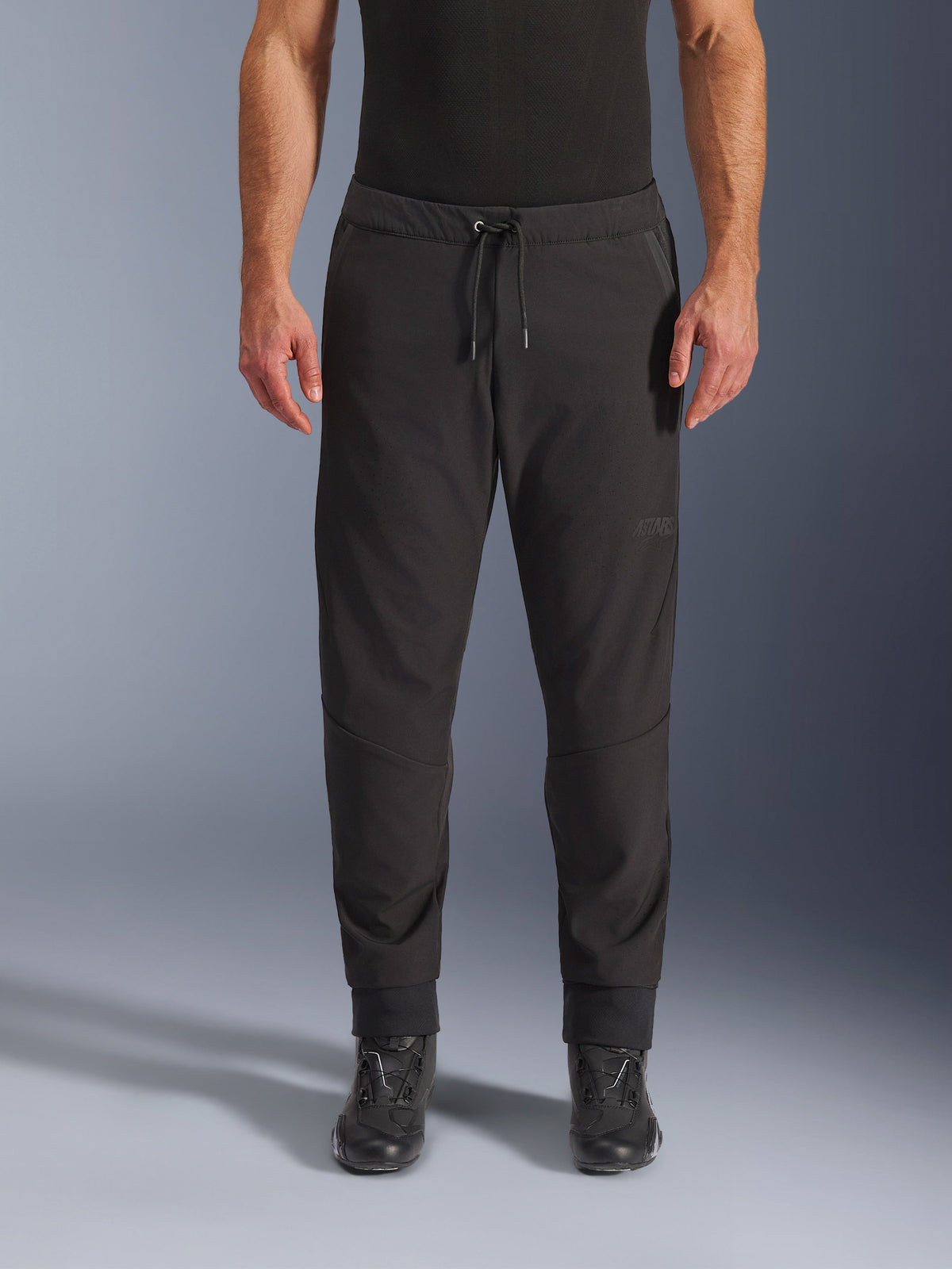 Alpinestars Aeroshell Airflow Pants, svarta motorcykelbyxor, vy framifrån bärs med svarta MC-stövlar, har dragsko i midjan och avsmalnande ankelmuddar, lätt softshell-design med subtil ton-i-ton-branding.