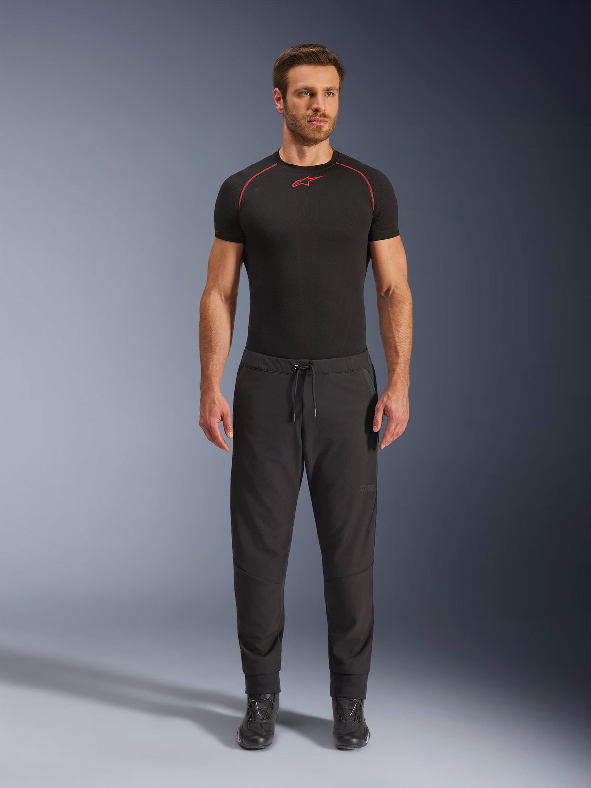 Modell som bär Alpinestars Aeroshell Airflow-byxor i svart, kombinerat med ett svart kortärmat underställ och svarta motorcykelstövlar. Dessa lätta softshell-motorcykelbyxor har dragsko i midjan och elastiska muddar, designade för sport- och touringförare i varma väderförhållanden.