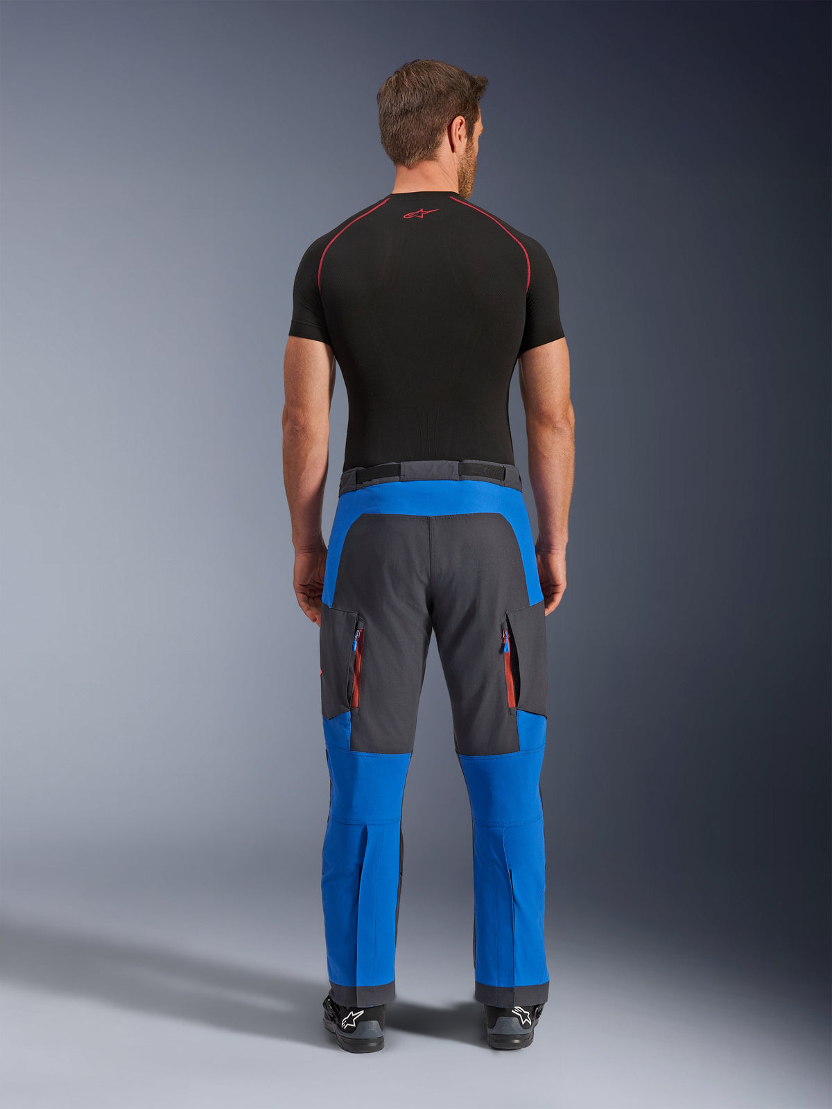 Alpinestars Maxdura Dual byxor i Royal Blue och Deep Gray, adventure-motorcykelbyxor sedda bakifrån, cargofickor med dragkedja och orangea detaljer, design för att bäras över stövlar, bärs med ett svart kortärmat underställ
