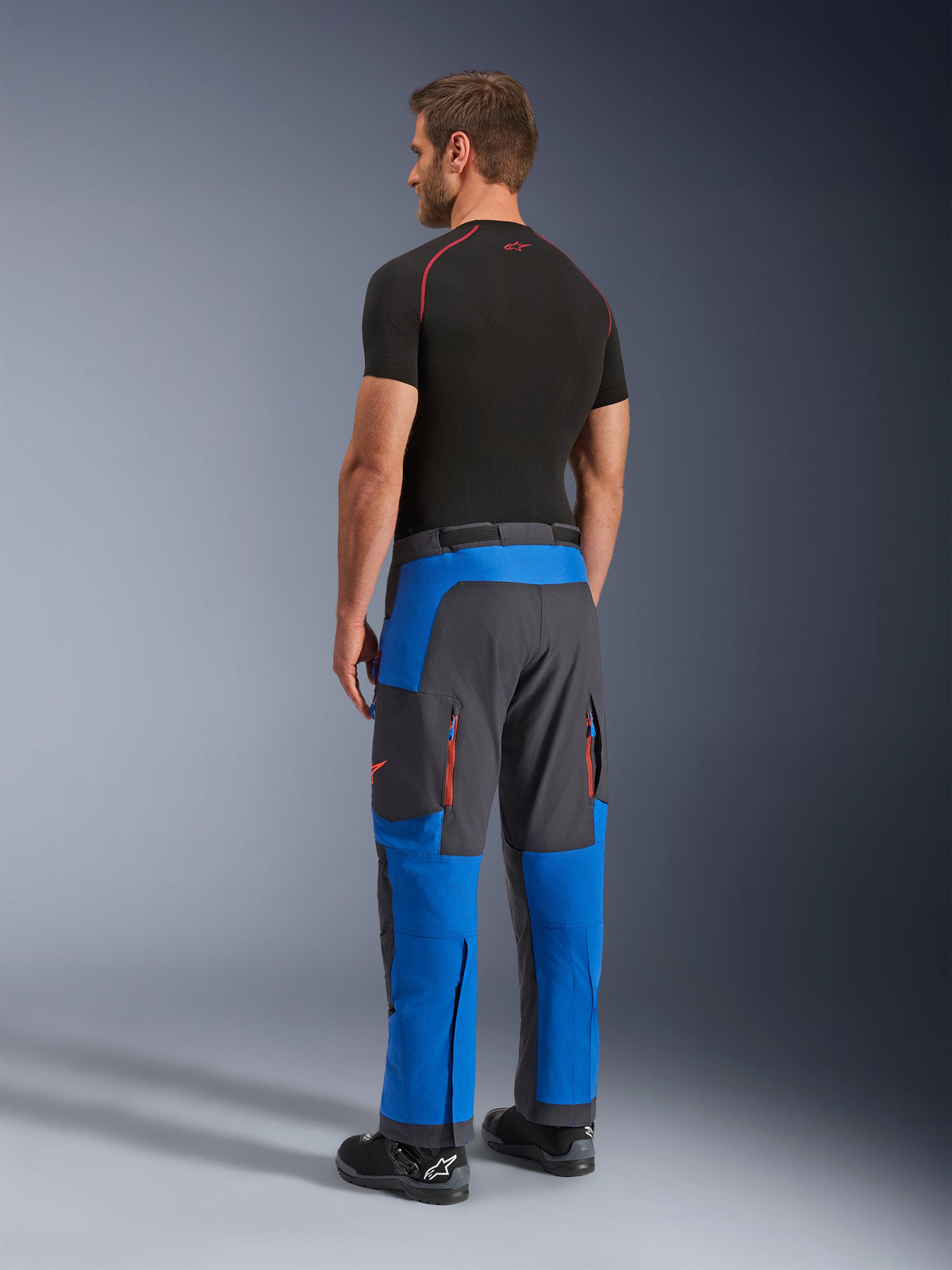 Alpinestars Maxdura Dual byxor, äventyrsbyxor för motorcykel, kungsblå och mörkgrå, sedda bakifrån på en modell, orangea ventilationsöppningar med dragkedja, passform över stövlar, slitstarkt stretchmaterial