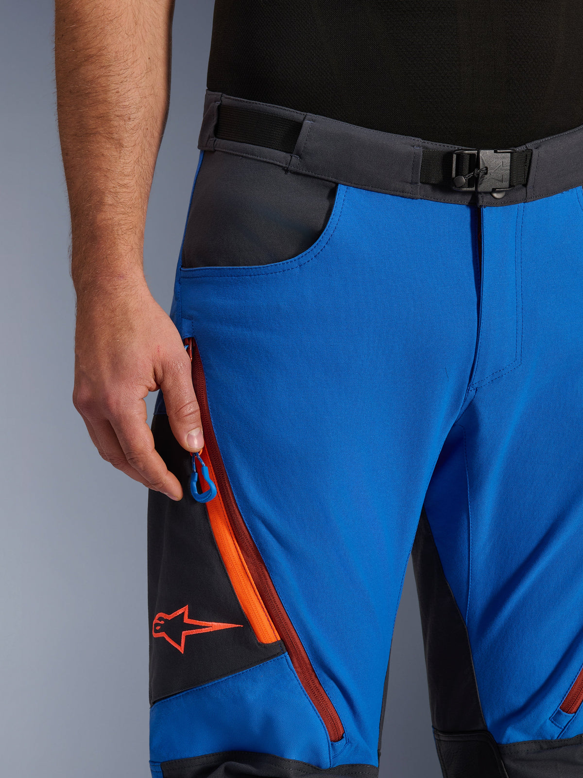 Alpinestars Maxdura Dual byxor i kungsblå och mörkgrå, närbild av midjebandet med magnetspänne, benficka med dragkedja med orangea detaljer och röd Alpinestars-logotyp, teknisk MC-utrustning för äventyrsåkning