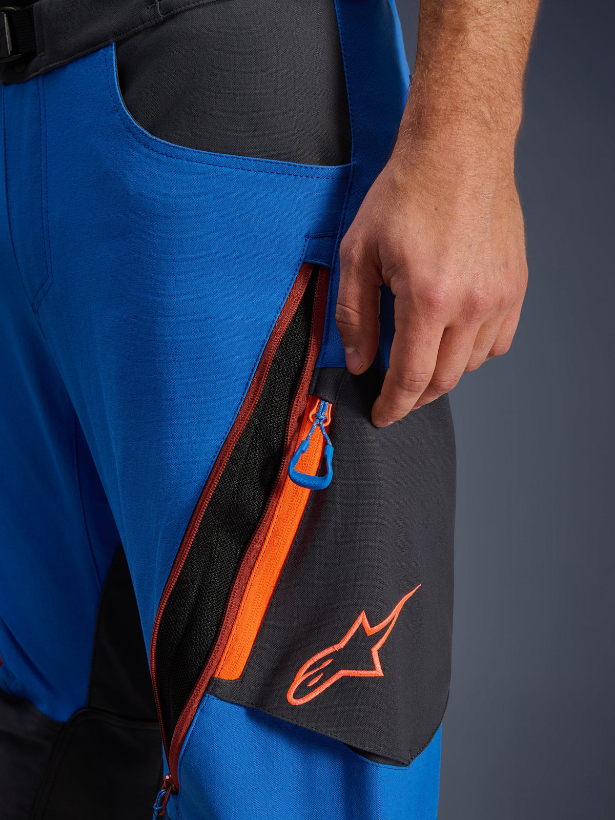 Alpinestars Maxdura Dual-byxor detalj, Royal Blue och Deep Gray äventyrs-mc-byxor, närbild på lårventilation med dragkedja med orangea accenter och blå dragflik, orange Alpinestars-logotyp på svart panel, hållbar konstruktion i stretch-polyamid