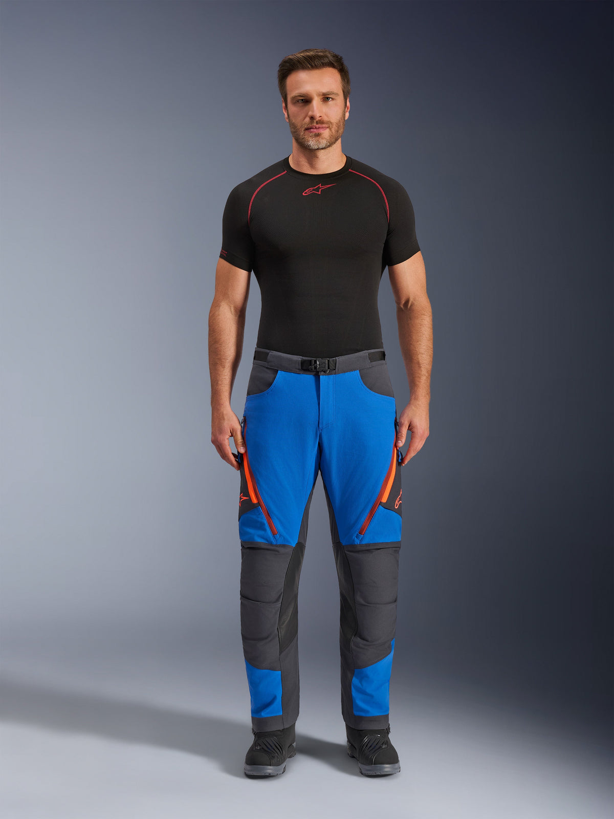 Alpinestars Maxdura Dual Pants, äventyrsmotorcykelbyxor, kungsblå och mörkgrå, orange dragkedjedetaljer, bärs med ett svart Alpinestars underställ och svarta stövlar, med förstärkta knäpaneler och en teknisk offroad-design