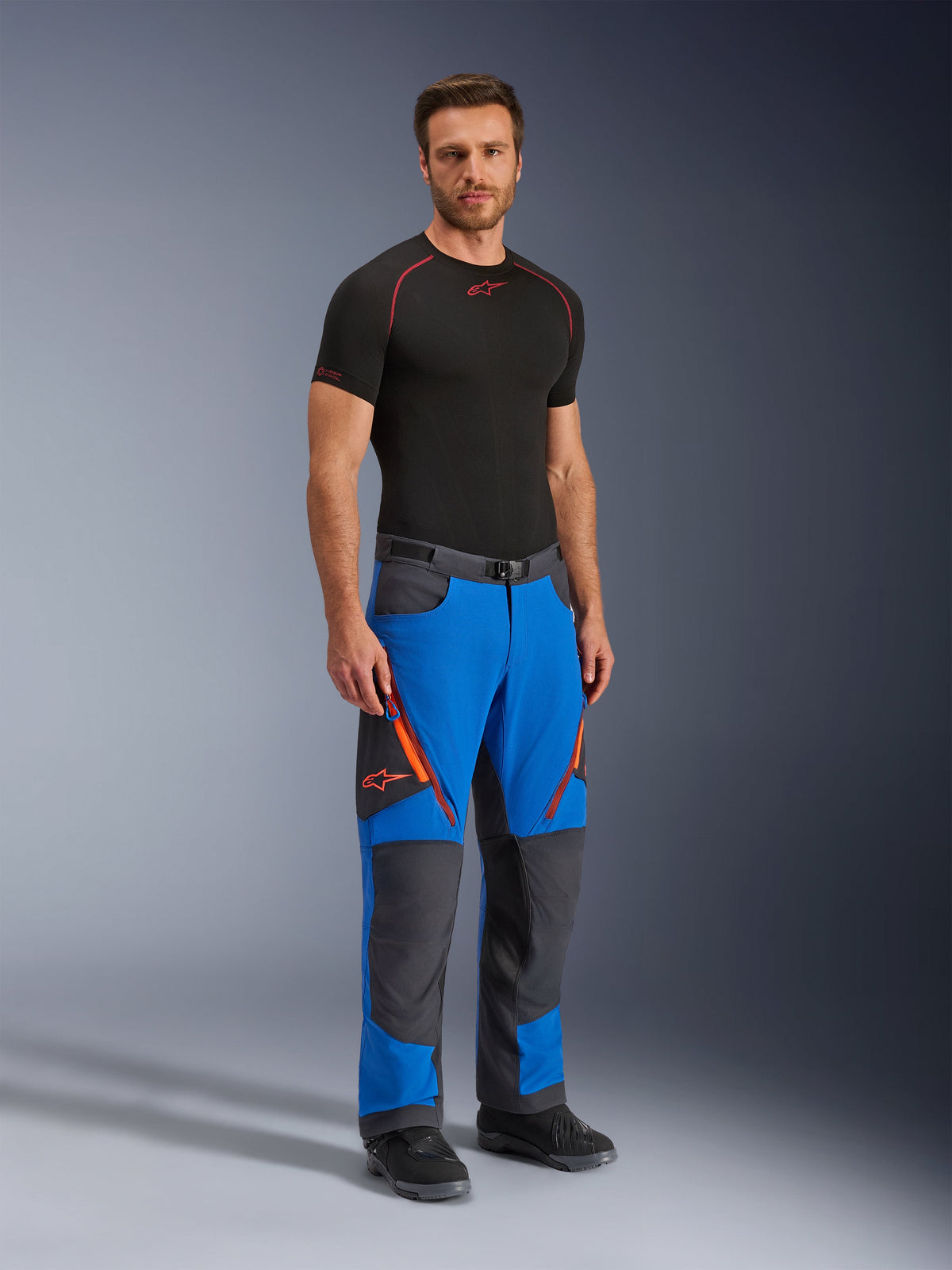 Alpinestars Maxdura Dual Pants, äventyrsmotorcykelbyxor, kungsblå och mörkgrå, med orangea dragkedjedetaljer och logotyper, bärs av en modell med svart underställ och stövlar, teknisk design med förstärkta paneler för dual-sport-körning