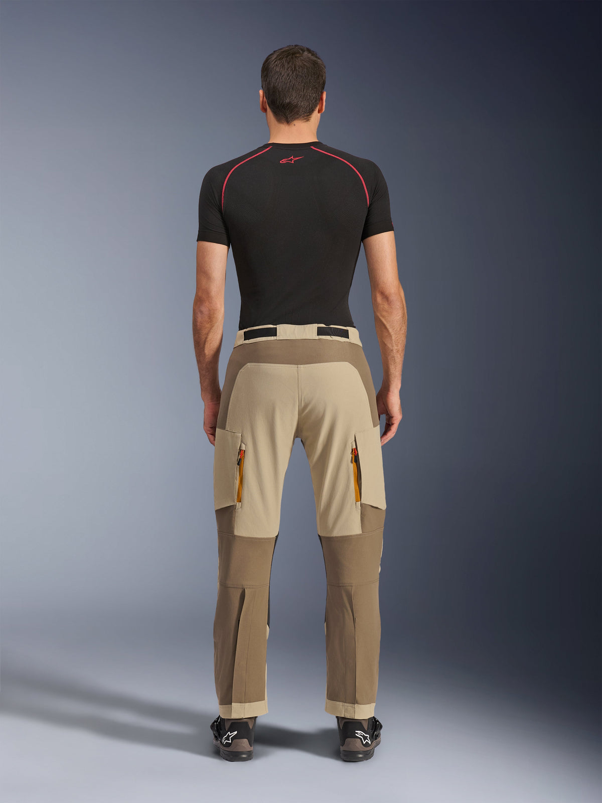 Alpinestars Maxdura Dual-byxor i Walnut Aluminum, sedda bakifrån, tvåtonad brun och beige design, äventyrsbyxor för motorcykelkörning med dragkedjeförsedda cargofickor och förstärkt säte, bärs med en svart kompressionströja och motorcykelstövlar.
