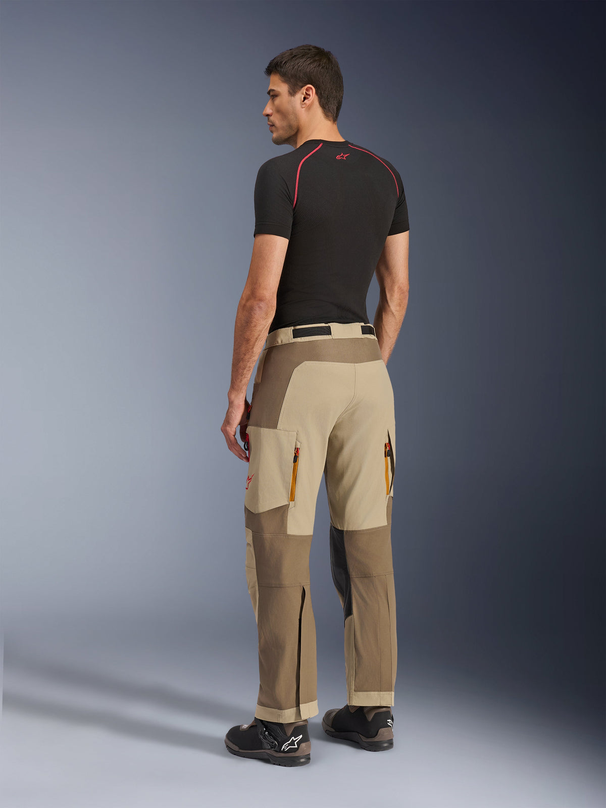 Alpinestars Maxdura Dual-byxor, äventyrs- och dual-sportbyxor, Walnut Aluminum, baksida bärs av en modell med svart underställ och mc-stövlar, med flerpanelskonstruktion, bakre ventilationsöppningar med dragkedja och förstärkta överlager.
