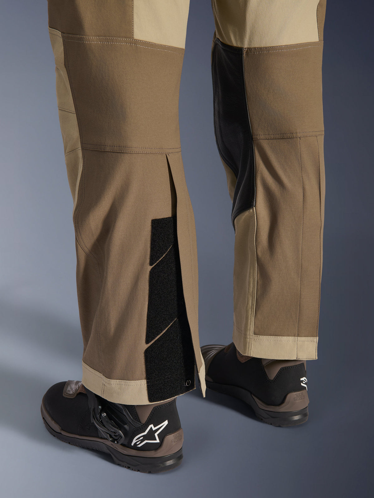 Alpinestars Maxdura Dual Pants i Walnut Aluminum, detaljerad vy av underben och nederkant med en over-the-boot-design, justerbara kardborrestängningar och förstärkta innerpaneler, matchade med svarta motorcykelstövlar.