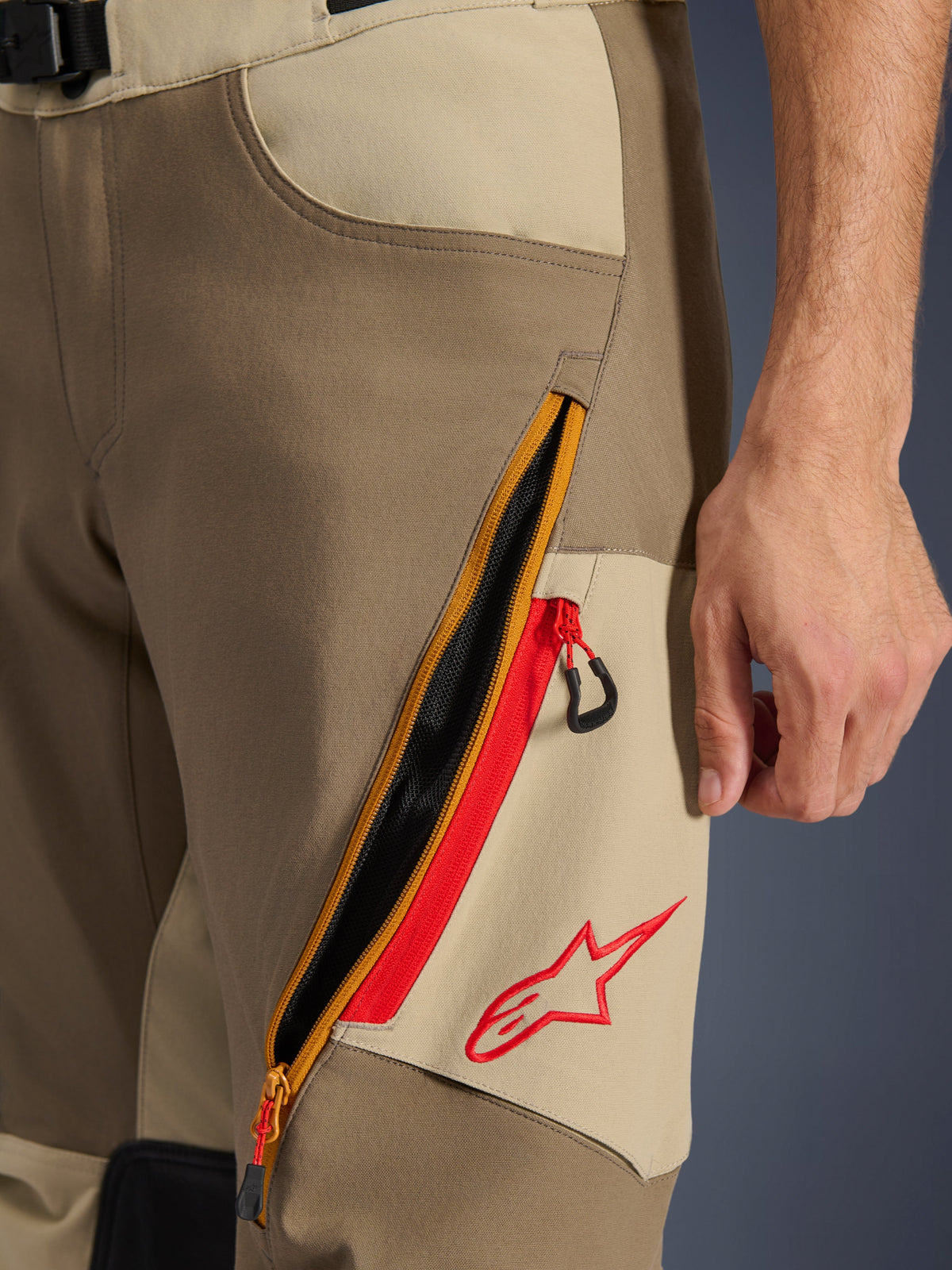 Alpinestars Maxdura Dual-byxor, Valnöt och Aluminium, närbild på lårdetalj med ett stort diagonalt ventilationssystem med dragkedja och röda accenter, röd Alpinestars-logotyp på en beige panel, äventyrsmotorcykelutrustning