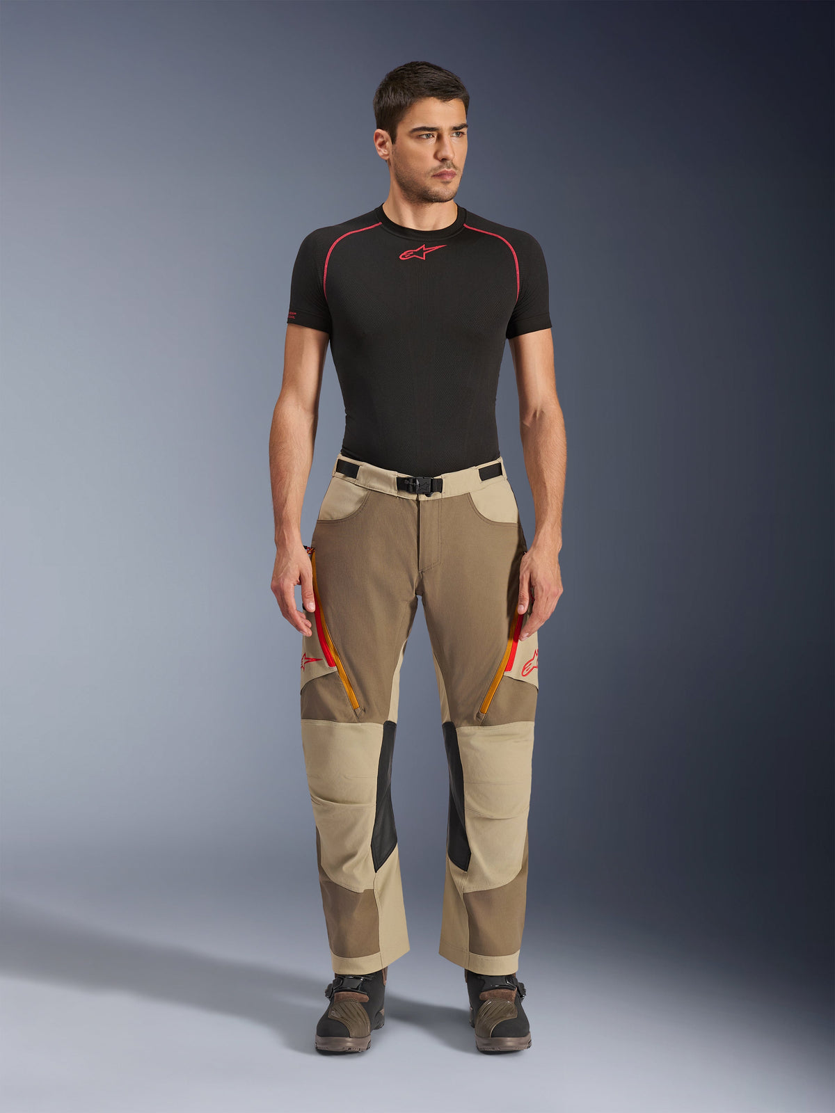 Alpinestars Maxdura Dual byxor, äventyrs- och dual-sport-byxor, Walnut Aluminum, bärs med en svart kortärmad kompressionströja och äventyrsstövlar, med orangea ventilationsöppningar med dragkedja och röda logotyper på låren, svart midjebälte med spänne, designade för att bäras över stöveln för motorcykelkörning.