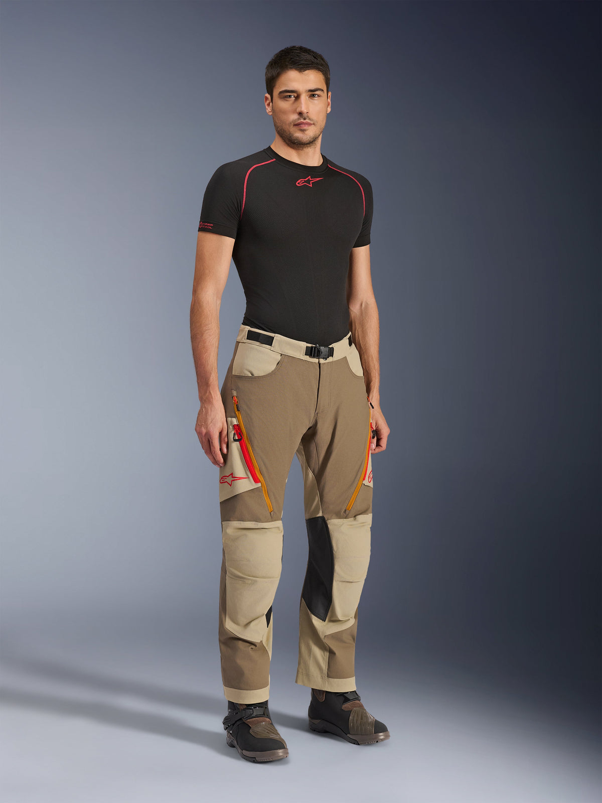 Alpinestars Maxdura Dual-byxor i Walnut Aluminum, bruna och beiga äventyrsmotorcykelbyxor med orangea dragkedjor och knäpaneler i svart läder, bärs med en svart baslager-t-shirt och äventyrsstövlar, helkroppsbild framifrån