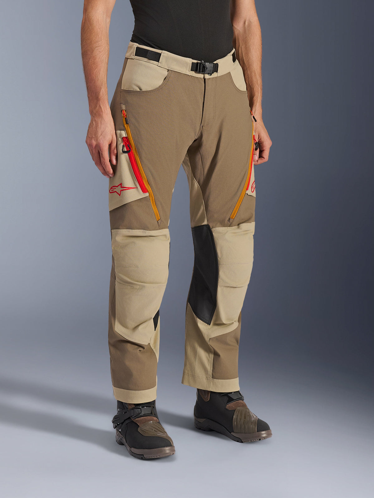 Alpinestars Maxdura Dual Pants, Walnut Aluminum bruna och khakifärgade äventyrsbyxor, med orangea och röda ventilationsöppningar med dragkedja på låren, förstärkta innerpaneler i svart läder vid knäna och röd Astars-logotyp, bärs med tekniska stövlar.