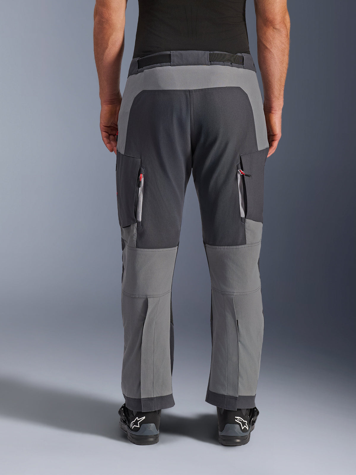 Alpinestars Maxdura Dual byxor, äventyrsmotorcykelbyxor, mörkgrå och djupgrå, vy bakifrån som framhäver förstärkta sätes- och knäpaneler, ventilationsöppningar med dragkedja på låren, bärs med svarta Alpinestars-stövlar, robust konstruktion i stretch-polyamid för dual-sport-körning