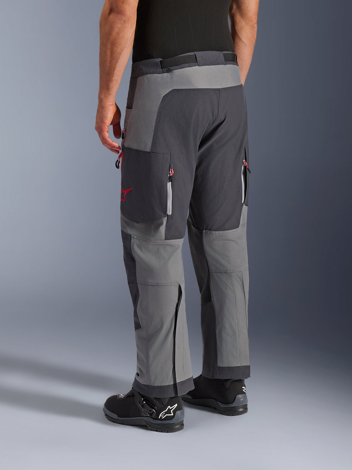 Alpinestars Maxdura Dual byxor, äventyrsbyxor för motorcykel, mörkgrå och djupgrå, vy bakifrån med flerpanelskonstruktion, röda dragkedjedetaljer och stjärnlogotyp, kombinerade med svarta Alpinestars-stövlar, designade för dual-sport-prestanda