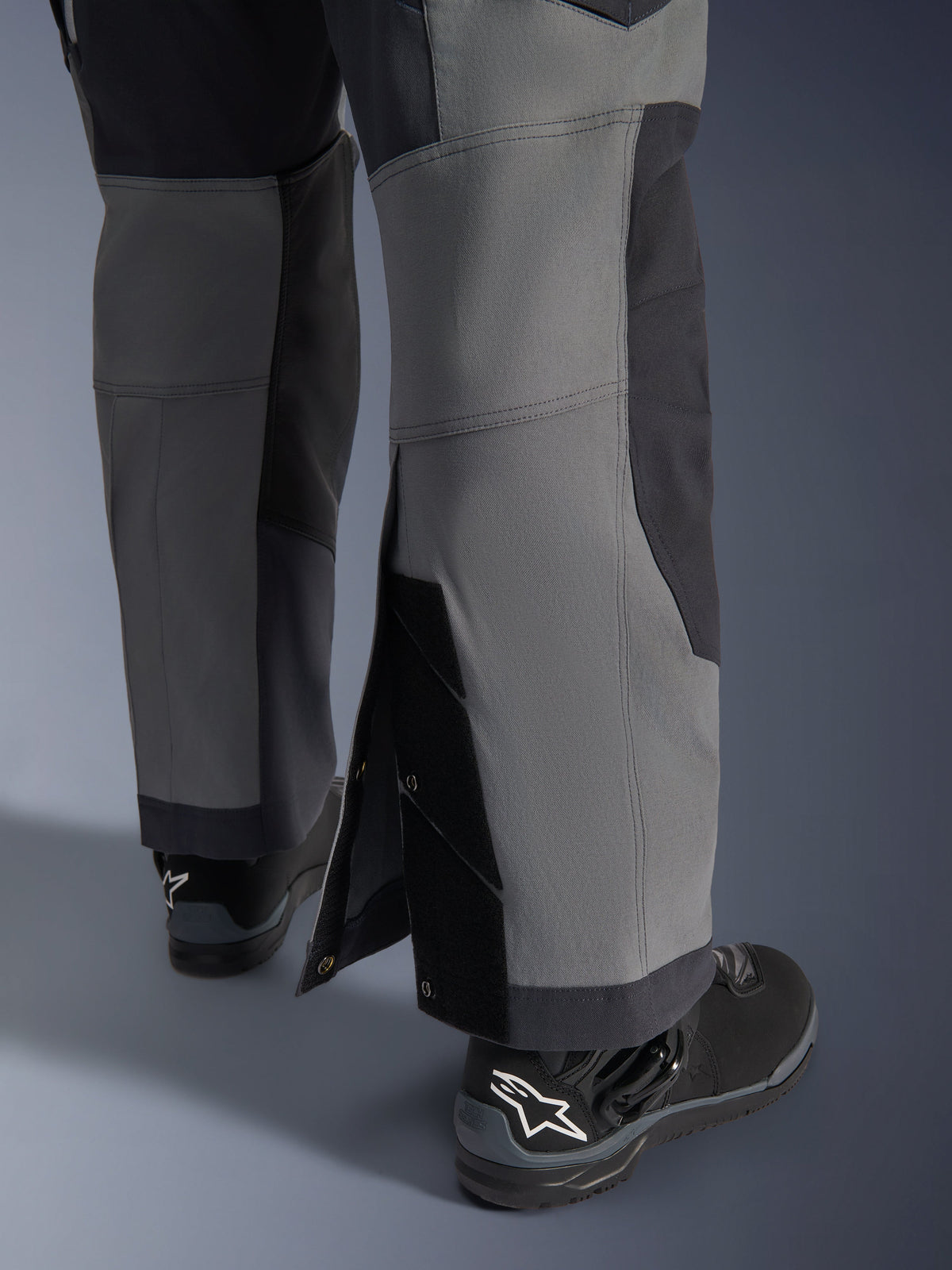 Alpinestars Maxdura Dual-byxor, äventyrsmotorcykelbyxor, mörkgrå och djupgrå, närbild av justerbara benslut som bärs över stövlarna med tryckknappar och förstärkta paneler, bärs med svarta Alpinestars-stövlar, robust teknisk design för dual-sport-körning