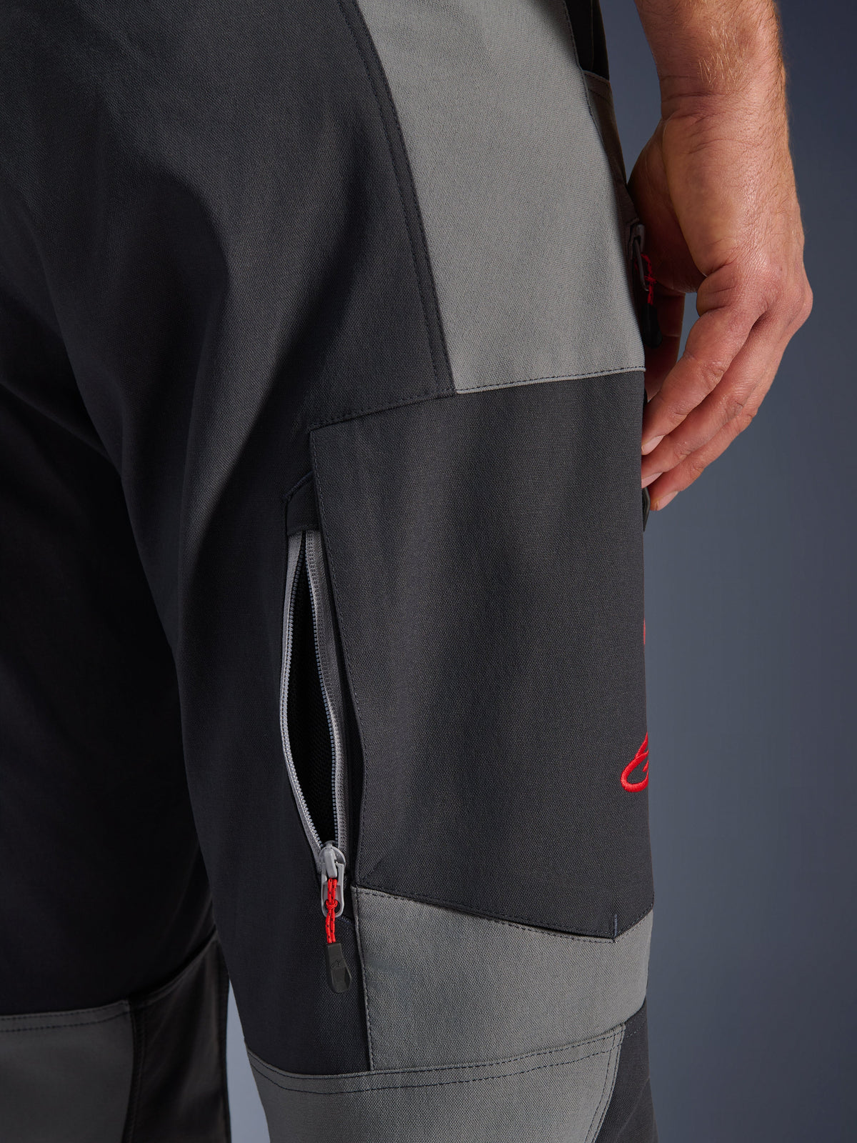Alpinestars Maxdura Dual byxor, äventyrsmotorcykelbyxor i mörkgrått och djupgrått, närbild på låret med en ventilationsöppning med dragkedja och röd broderad logotyp, hållbar konstruktion i stretch-polyamid.