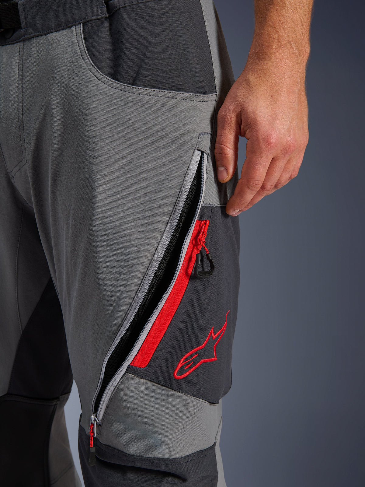 Alpinestars Maxdura Dual byxor, mörkgrå och djupgrå äventyrsbyxor för motorcykel, närbild på öppen lårventilation med dragkedja, röda detaljer och broderad röd stjärnlogotyp, teknisk stretchvävsdesign