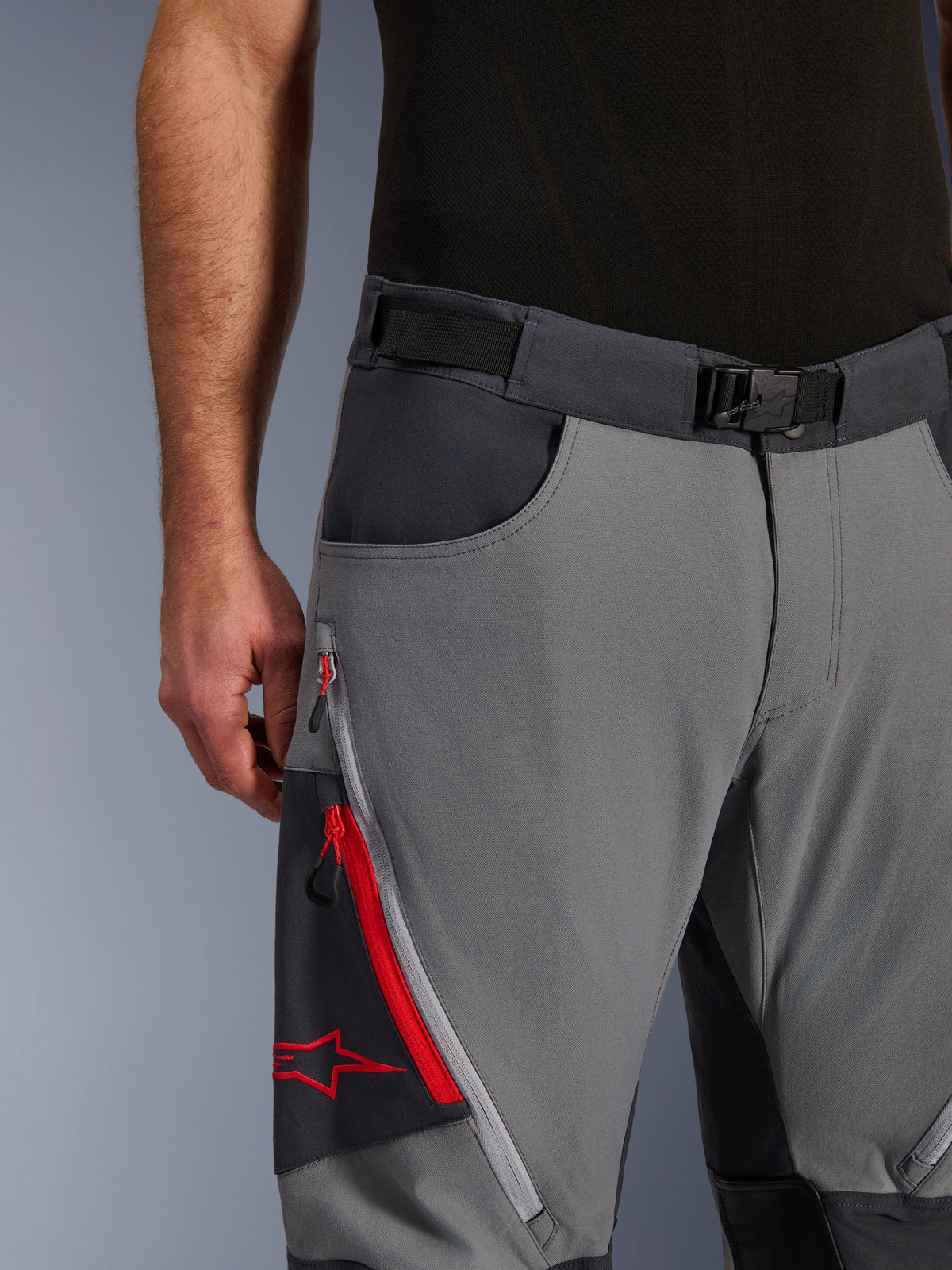 Alpinestars Maxdura Dual Pants, adventure-motorcykelbyxor, mörkgrå och djupgrå, närbild som visar justerbart midjebälte med magnetspänne, benficka med dragkedja med röda detaljer och Alpinestars-logotyp.