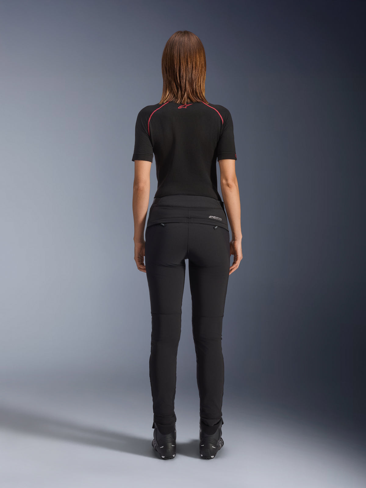 Alpinestars Alamosa dambyxor, svarta, vy bakifrån som visar en figurnära textilkonstruktion, bakfickor med dragkedja och reflekterande MO.ST.EQ.-logotyp på det bakre oket, bärs med ett svart kortärmat baslager och svarta motorcykelstövlar