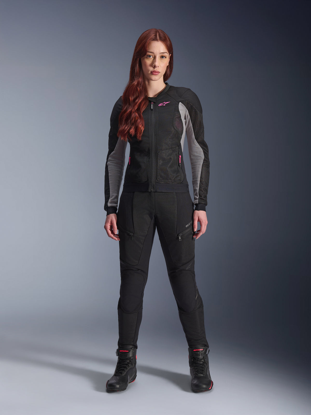 Alpinestars Stella Troop-Air-byxor, motorcykelbyxor för dam i svart, har meshpaneler och fickor med dragkedja, stylade med en matchande svart och grå meshjacka med rosa detaljer och svarta motorcykelstövlar, komplett outfit för urban motorcykelkörning.