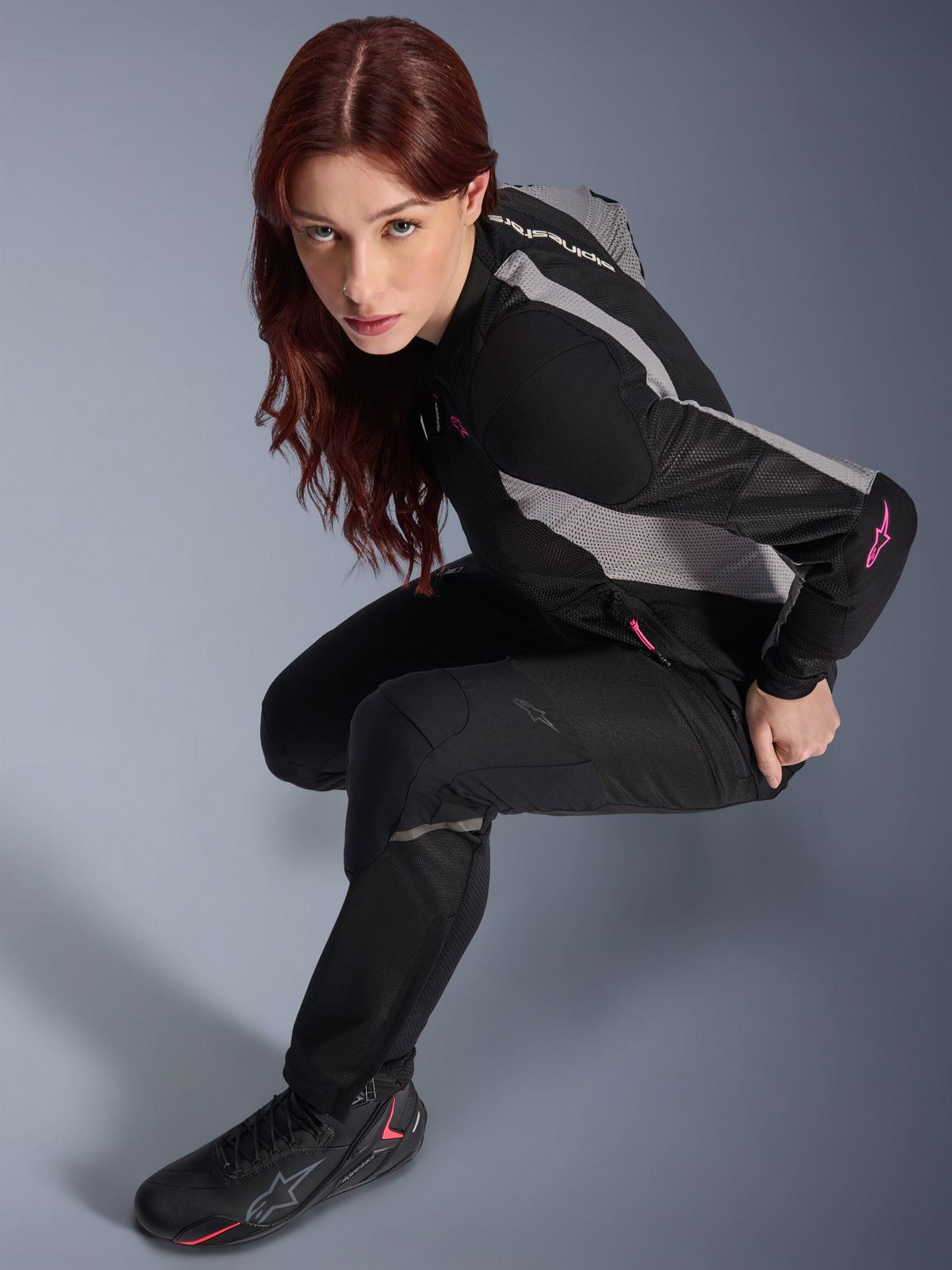 Kvinna som bär Alpinestars Stella Troop-Air byxor, svarta, lätta ventilerade textilbyxor, matchade med en svart och grå mesh-jacka med rosa detaljer och svarta mc-stövlar, studioporträtt, urban motorcykelutrustning