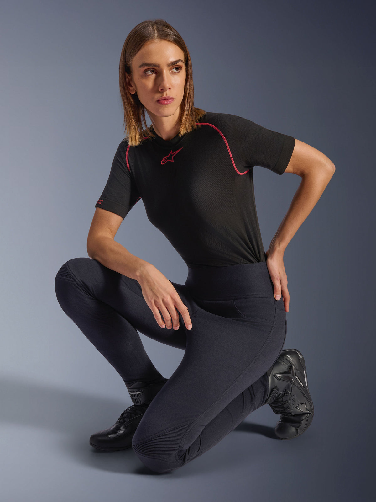 Kvinna som bär Alpinestars Stella Flex-AST Leggings, svarta motorcykelleggings med hög midja och smal passform med dragspelsknäpaneler, stylade med en svart kortärmad funktionst-shirt med rosa sömmar och mc-stövlar.