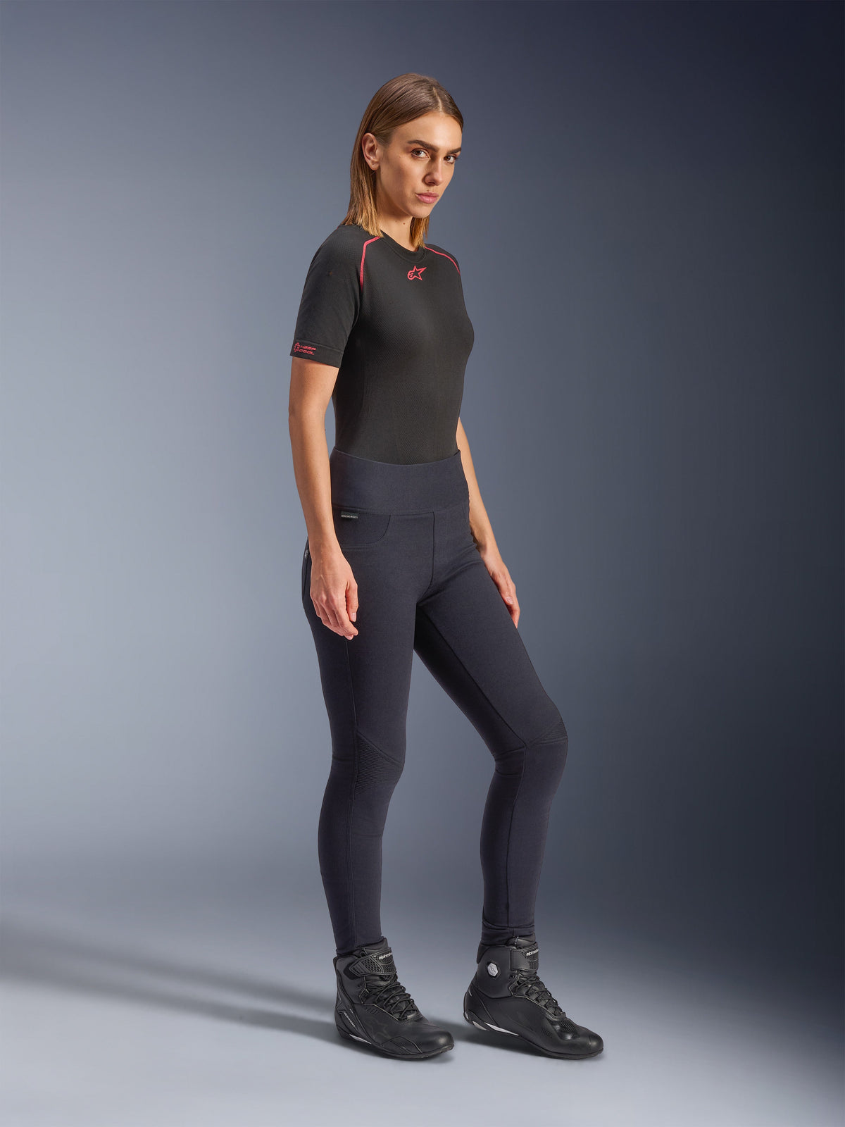 Modell som bär Alpinestars Stella Flex-AST Leggings för dam i svart, hög midja och smal passform med dragspelsknäpaneler, kombinerat med en svart kortärmad Alpinestars-topp och svarta motorcykelstövlar, helkroppsbild i studio.