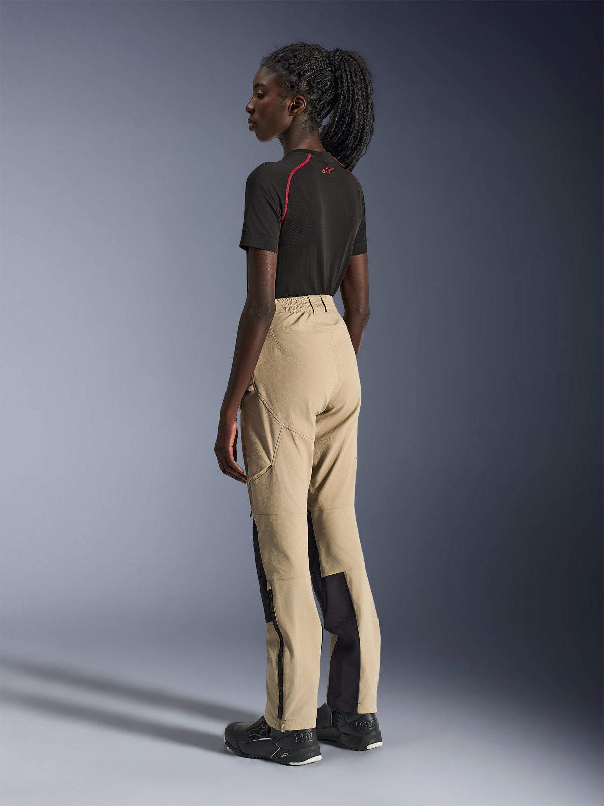 Alpinestars Stella Flex-AST Explorer Pants, mc-byxor för damer, sandfärgade och svarta, sidovy bakifrån på en modell kombinerat med en svart t-shirt och mc-stövlar, framhäver ripstop-förstärkningar och dragkedjeöppningar vid bensluten.