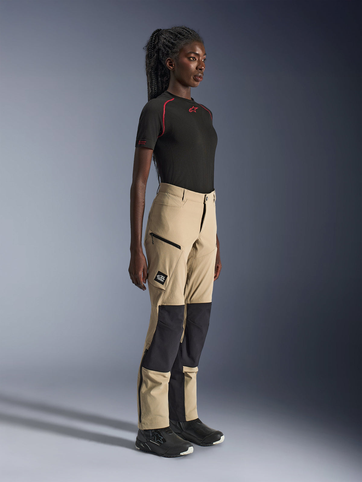 Alpinestars Stella Flex-AST Explorer dam-byxor, sandfärgade och svarta adventure touring-byxor, sidoprofil på en modell som bär en svart Alpinestars t-shirt och körstövlar, med ripstop-förstärkningar på knäna och cargofickor med dragkedja.