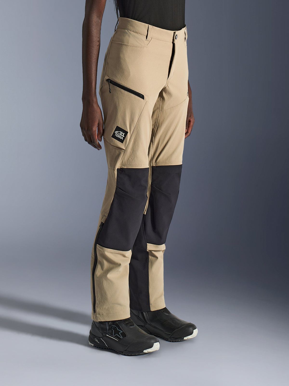 Kvinna som bär Alpinestars Stella Flex-AST Explorer Pants, sandfärgade och svarta äventyrsmotorcykelbyxor med svarta ripstop-knäpaneler, cargofickor med dragkedja, 'ALL TERRA' logotyp-patch och dragkedjor vid bensluten.