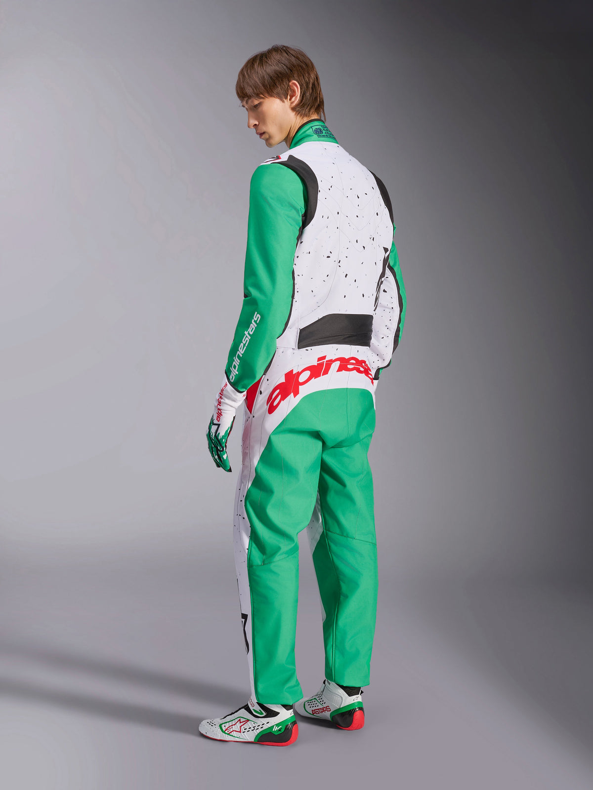 Alpinestars KMX-9 V3 Graphic 6-dräkt, kartingoverall, vit, grön och röd, modell sedd bakifrån, med gröna ärmar och nedre paneler, vit överkropp med svarta stänkdetaljer, framträdande röd Alpinestars-logotyp över ländryggen, matchad med vita och gröna kartingskor