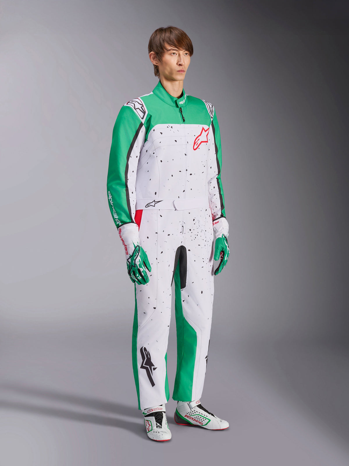 Alpinestars KMX-9 V3 Graphic 6-dräkt, kartingoverall, vit, grön och röd, bärs med matchande racinghandskar och skor, vit bas med gröna ärmar och svarta stänkmönster, röd Alpinestars-logotyp på bröstet, atletisk design i ett stycke för motorsport.