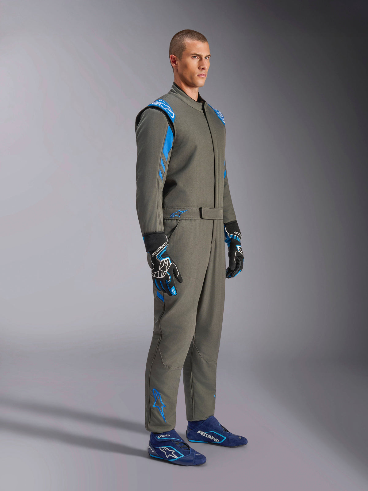 Alpinestars GP Race V3 Suit FIA i Anthracite Blue, racingoverall i ett stycke som bärs av en manlig modell, har klarblå accenter på axlar och anklar, kombinerad med matchande racinghandskar och blå racingskor, professionell motorsportutrustning.
