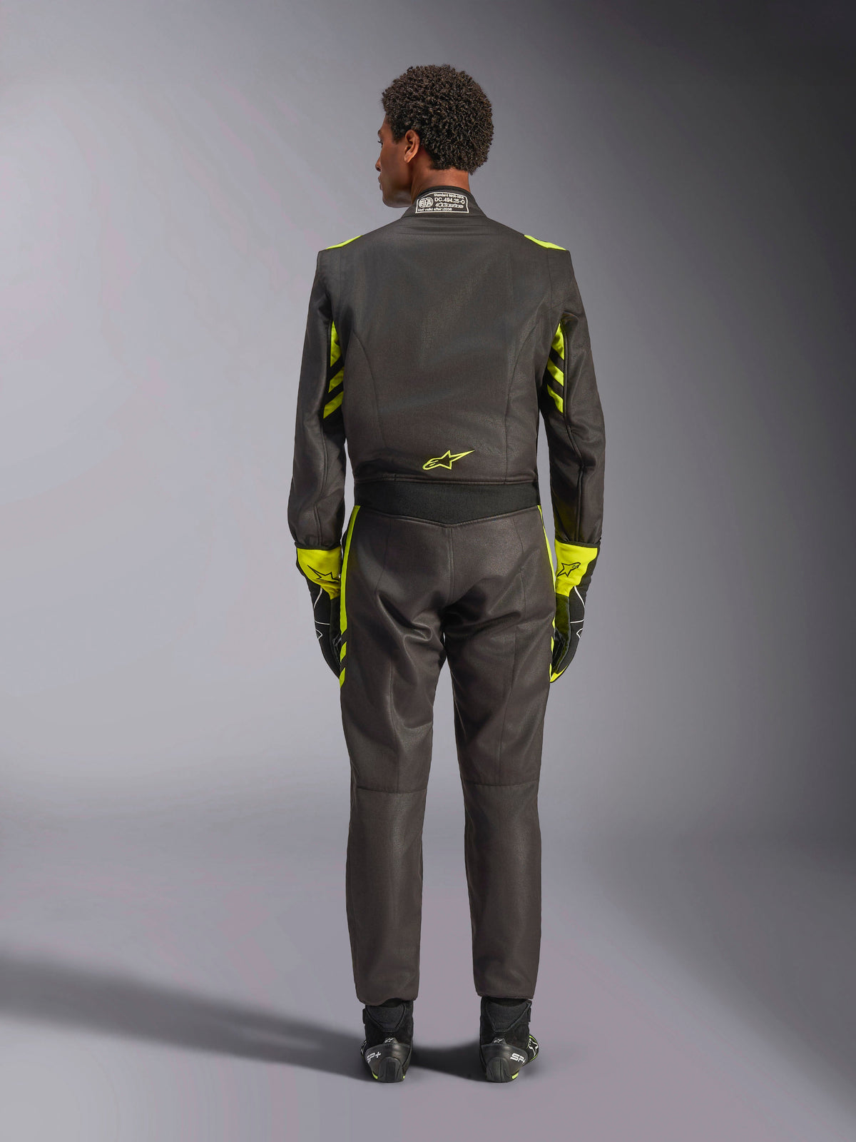 Alpinestars GP Race V3 Suit FIA, vy bakifrån, svart med neongula detaljer, med gula paneler på armarna och överkroppen, gul logotyp på nedre delen av ryggen, bärs med matchande racinghandskar och stövlar, professionell racingoverall för bilsport.