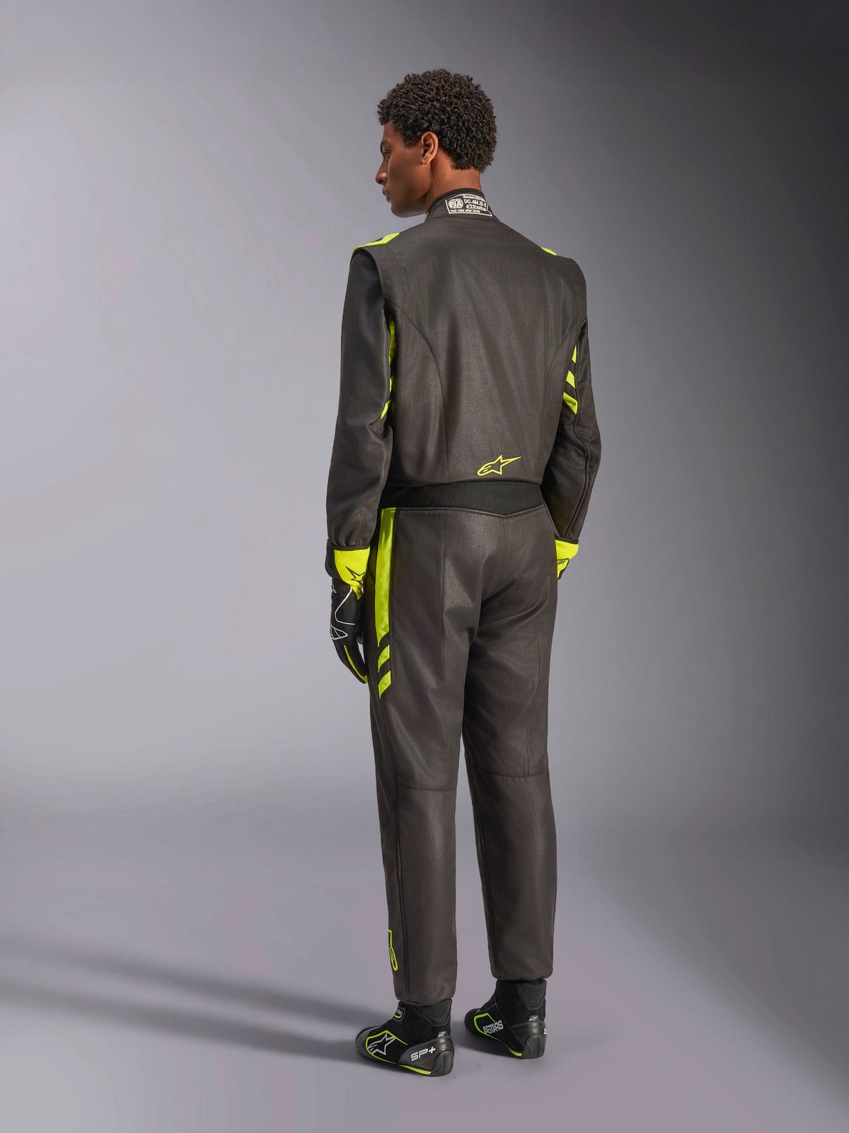 Baksida av Alpinestars GP Race V3 Suit FIA, svart med gula fluo-detaljer, bärs med matchande racinghandskar och racingstövlar, professionell bilsportutrustning med tvålagers aramidkonstruktion