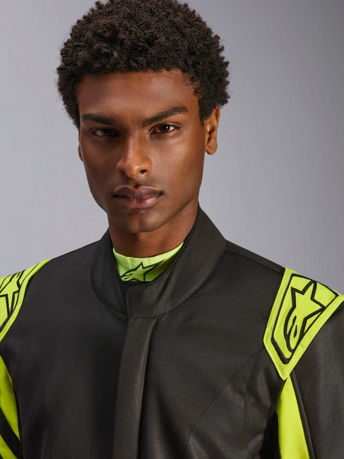 Närbild på en modell som bär Alpinestars GP Race V3 Suit FIA i Black Yellow Fluo, med gula extraktionsaxelremmar på axlarna med stjärnlogotyper, hög kragdesign, professionell bilsportutrustning.