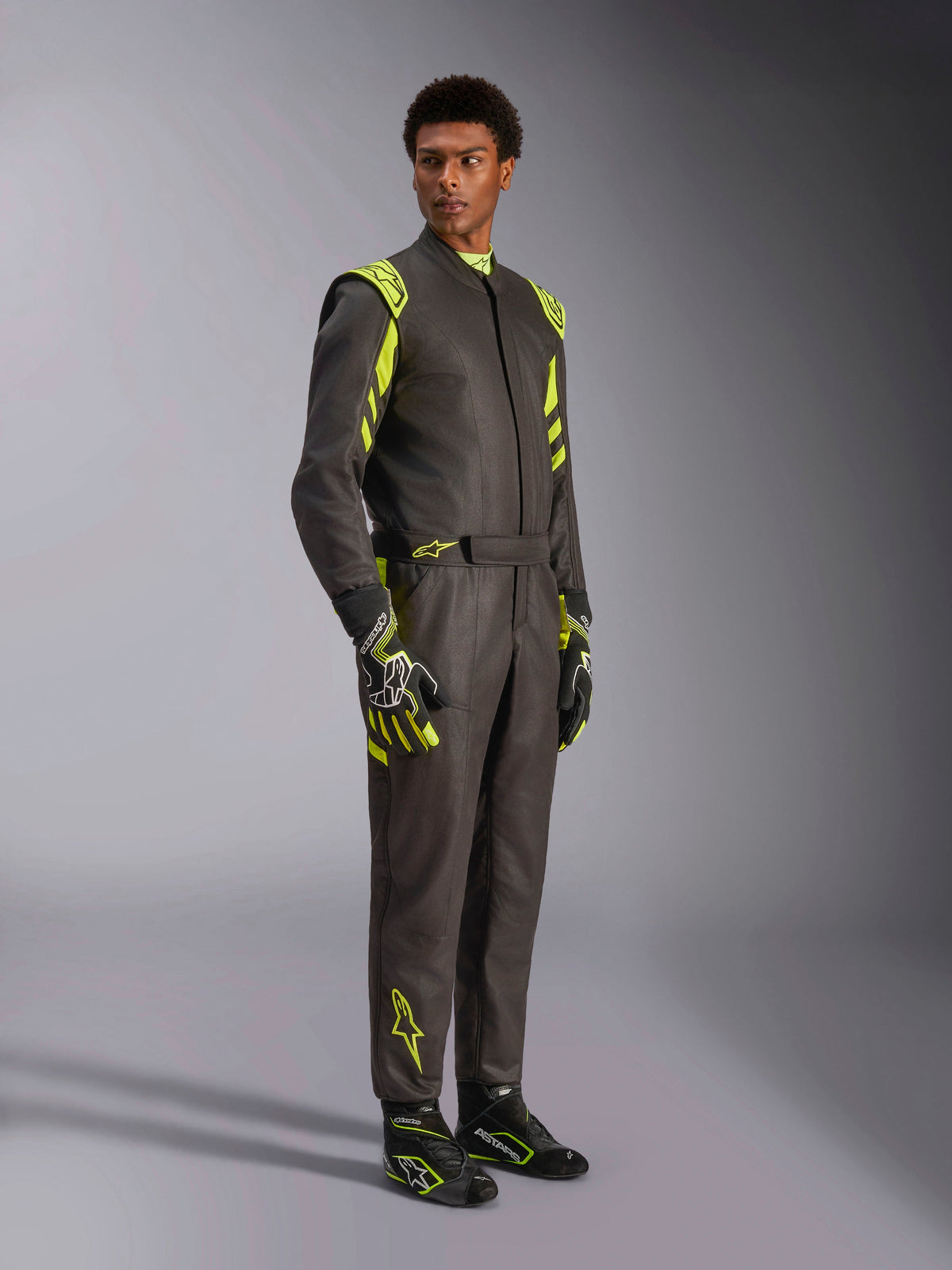 Alpinestars GP Race V3 Suit FIA, racingoverall för bilsport, svart och neongul, bärs av en modell med matchande racinghandskar och racingstövlar, iögonfallande neongula detaljer på axlar och ben, professionell motorsportutrustning