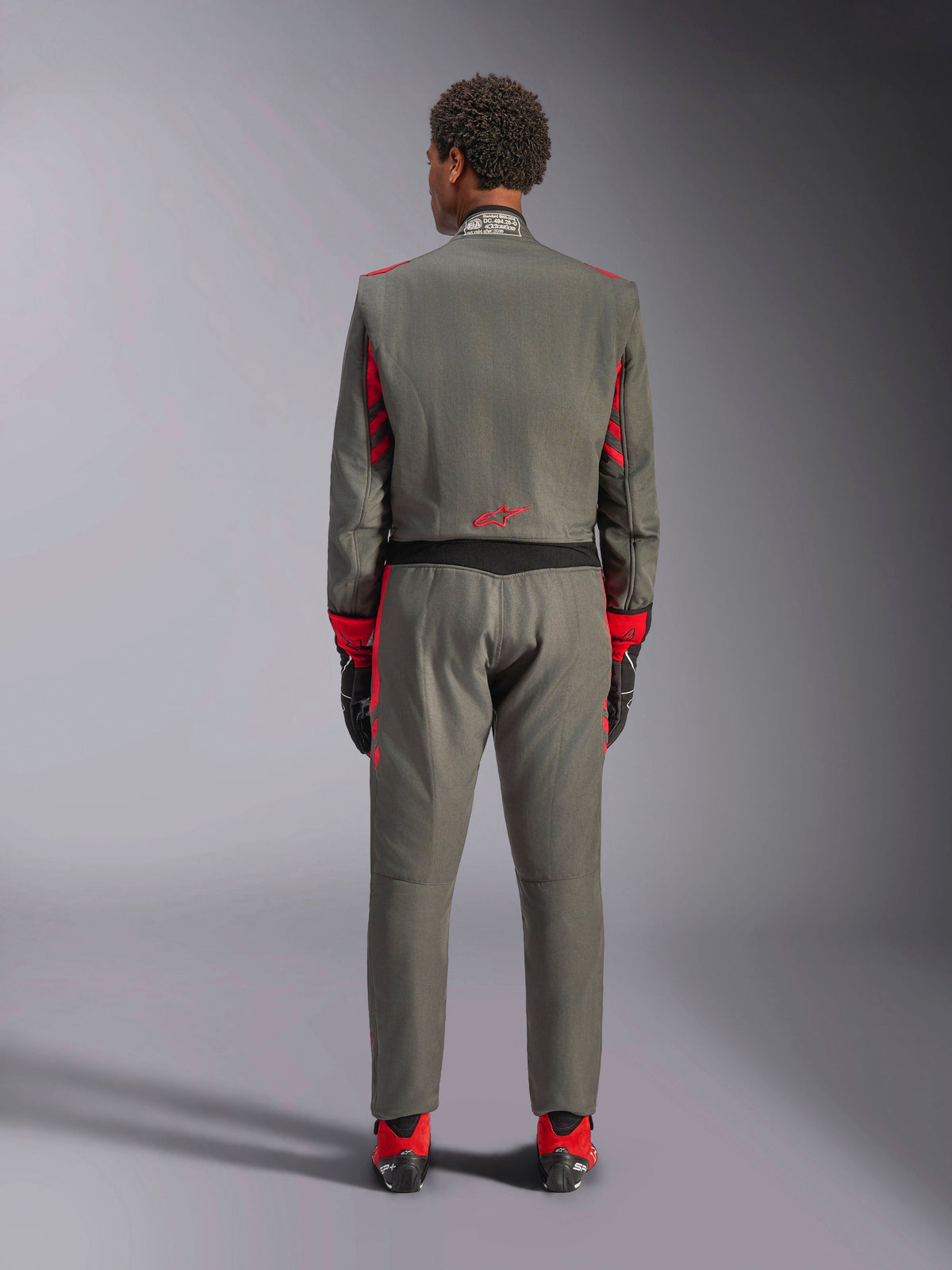 Bakifrån av en modell som bär Alpinestars GP Race V3 Suit i Anthracite Red, professionell bilracingdräkt med röda detaljer på ärmar och ben, kombinerad med röda racingstövlar och svarta handskar, teknisk motorsportutrustning i en studiomiljö.