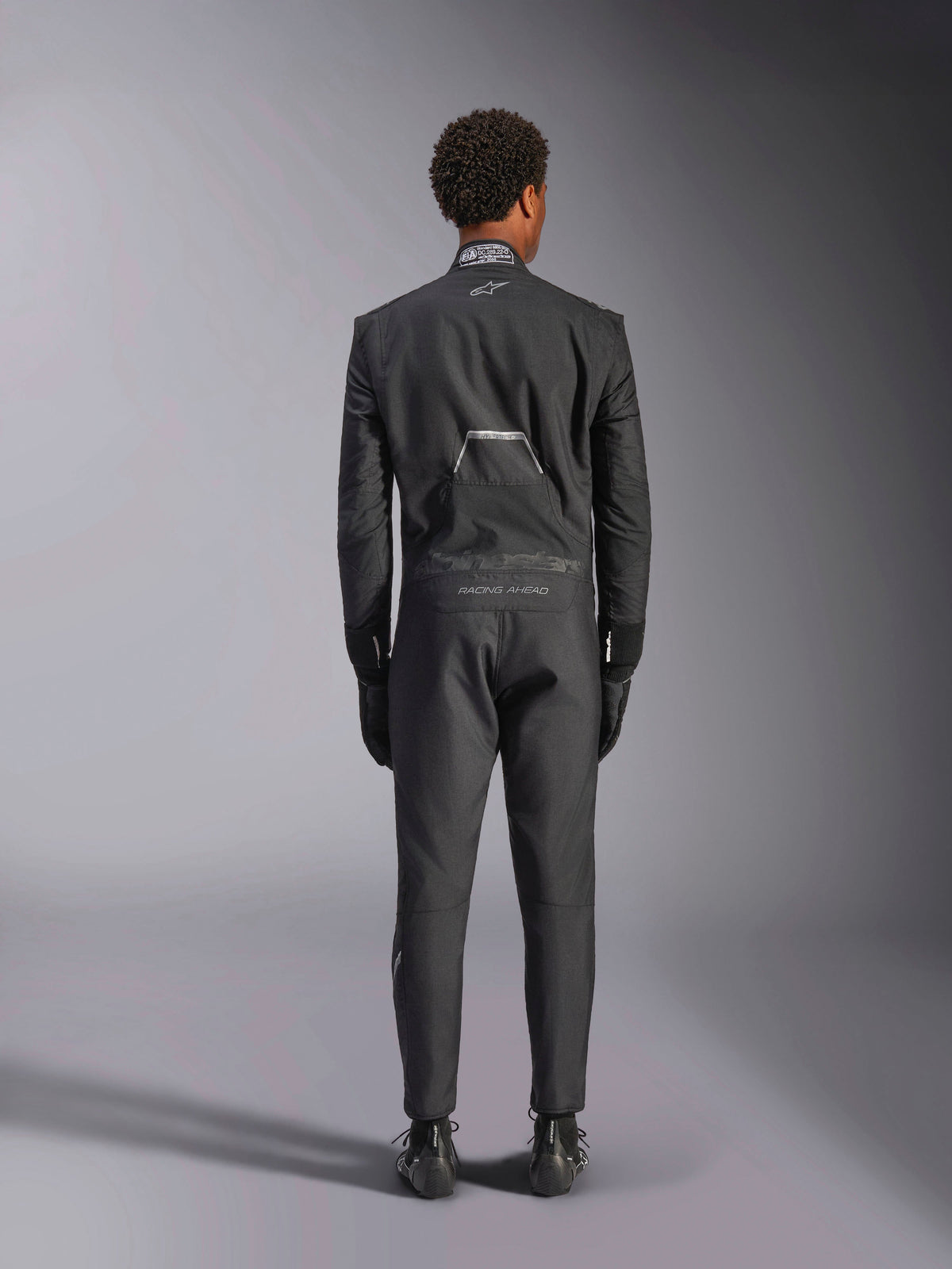 Alpinestars Hypertech V3 Suit, Black Dark Shadow, baksida av en professionell racingoverall i ett stycke för bilracing, med FIA-certifieringsetikett på kragen och "Alpinestars" och "RACING AHEAD"-märkning på ländryggen, bärs med matchande svarta racinghandskar och stövlar.