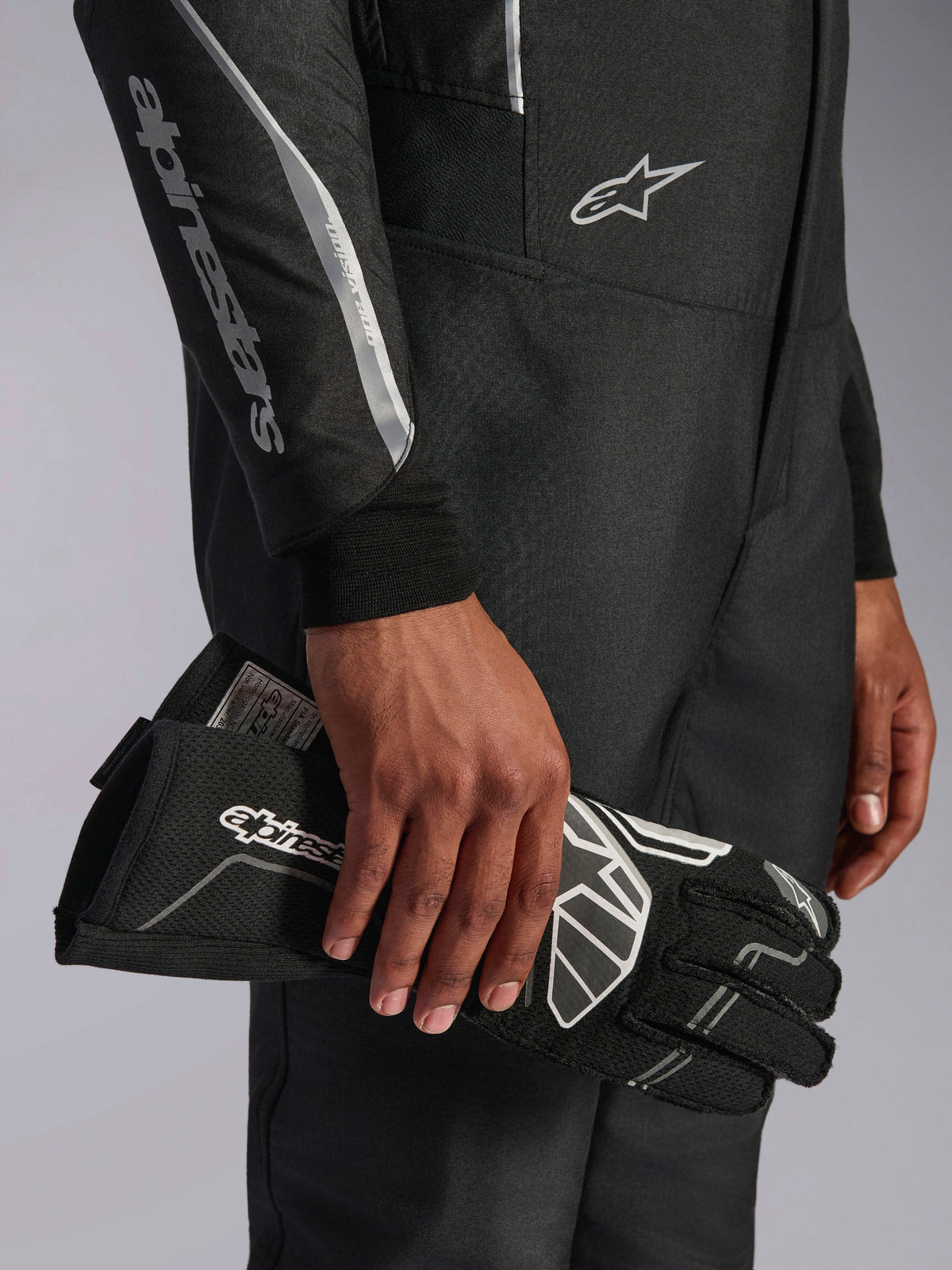 Alpinestars Hypertech V3-dräkt i Black Dark Shadow, närbild på ärm och torso med Alpinestars-branding i silver, en hand som håller en svart racinghandske med vita detaljer, professionell racingutrustning