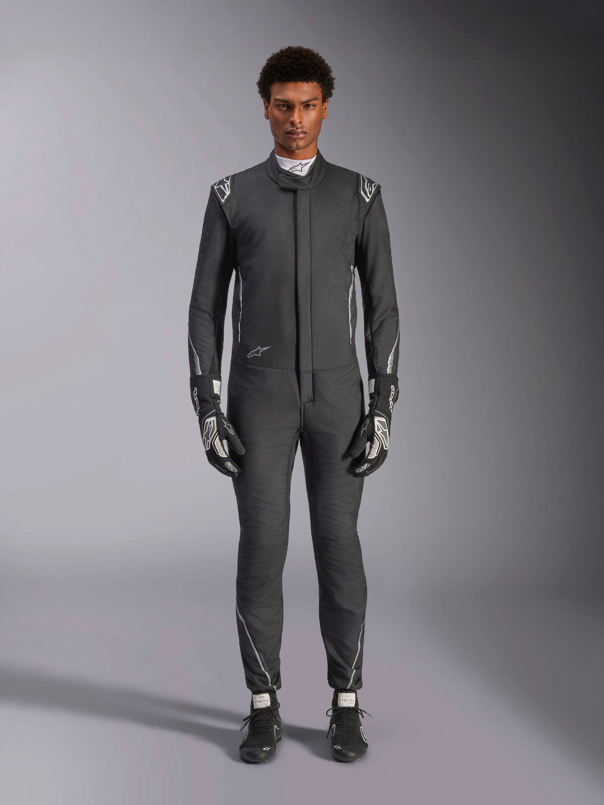 Alpinestars Hypertech V3 Suit, professionell racingoverall för bilsport, Black och Dark Shadow, bärs av en modell med matchande racinghandskar och skor, slimmad atletisk passform med silverdetaljer och logotyper, högpresterande motorsportutrustning