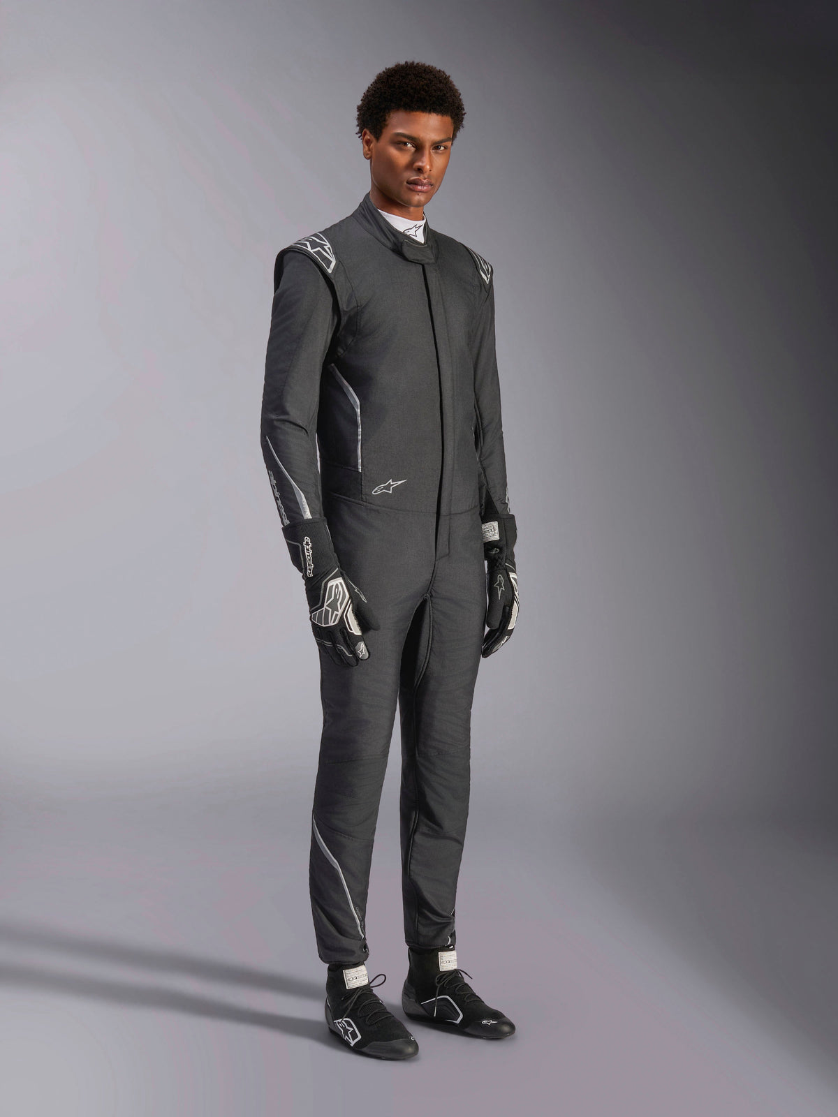 Alpinestars Hypertech V3-dräkt, racingoverall, svart och Dark Shadow, bärs av en manlig modell med matchande svarta handskar och racingskor, atletisk slim fit, har Alpinestars-logotyper och ljusgrå accenter, professionell skyddsutrustning för motorsport