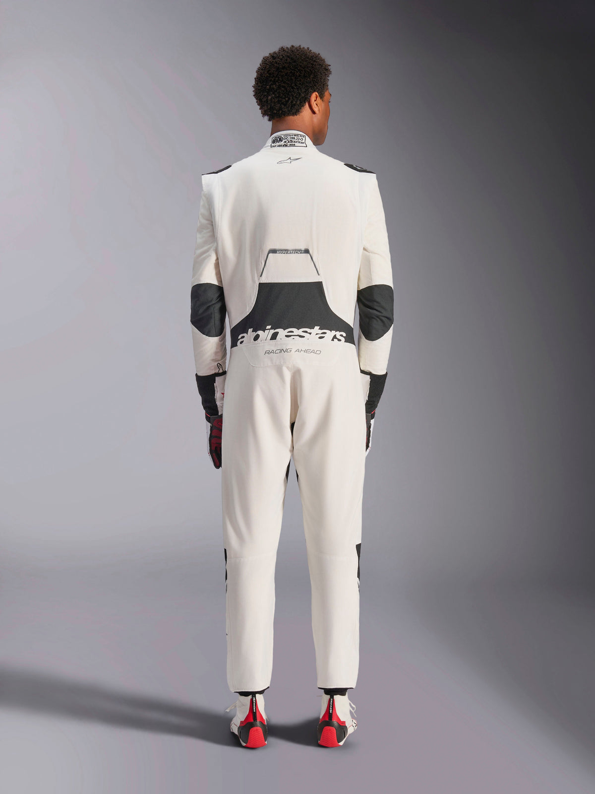 Bakifrån av en modell som bär Alpinestars Hypertech V3-dräkten i vitt, svart och silver, som visar den stora svarta stretchpanelen i ländryggen med 'alpinestars'- och 'RACING AHEAD'-logotyper, svarta armbågsinsatser, FIA-certifieringsetikett på kragen och en atletisk passform designad för professionell racing.